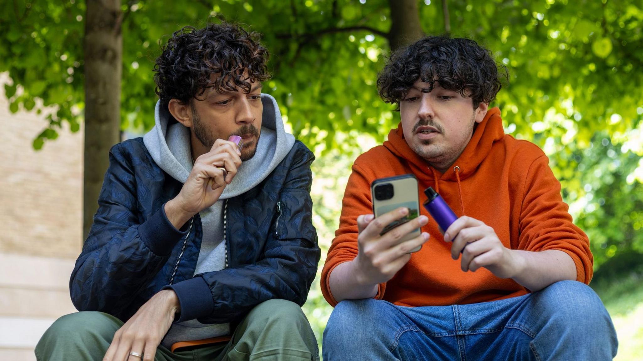 Two young men sit outside vaping. They are chatting, while looking at a mobile phone. 