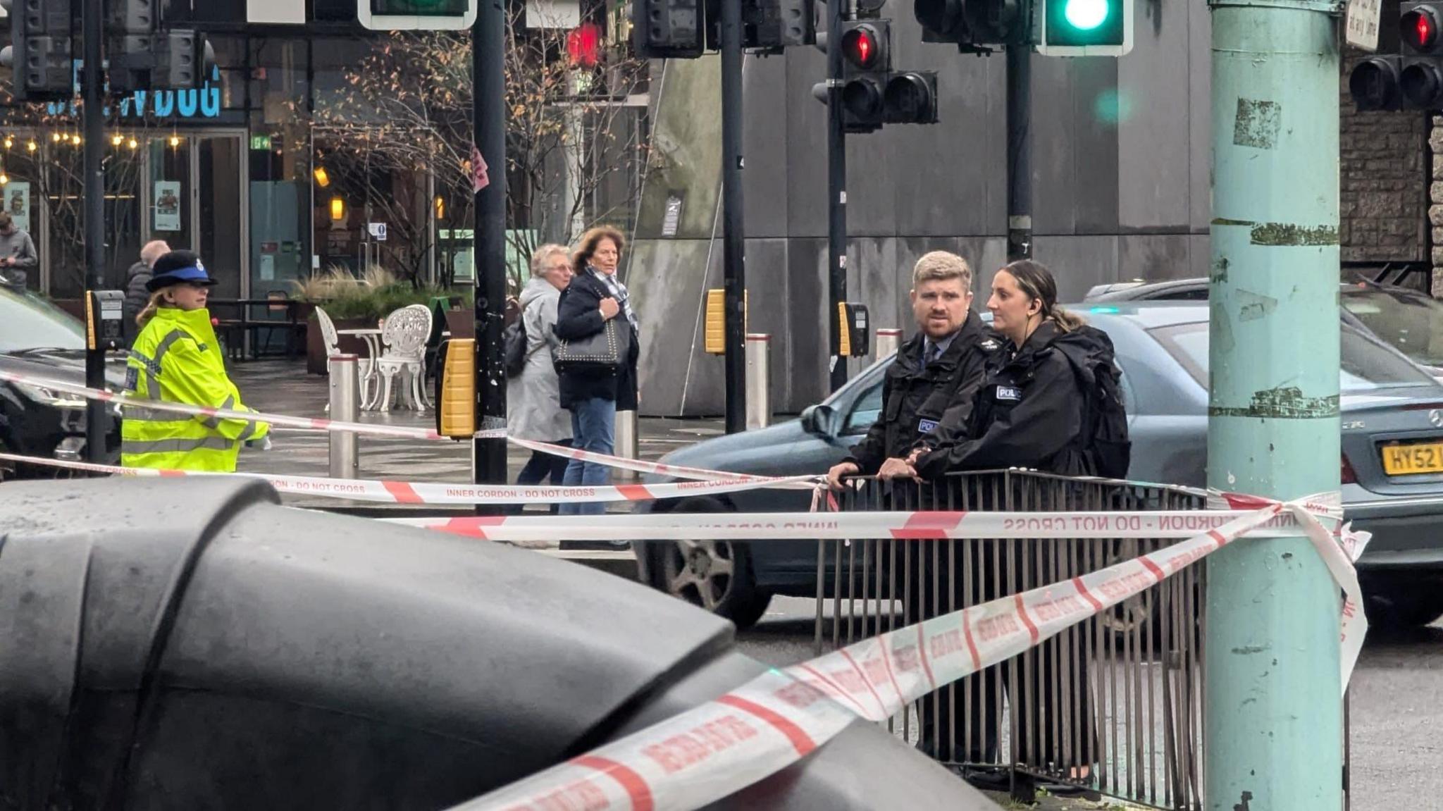 Two police officers stood against a fence on the right with a silver car driving behind. There is another police officer wearing a yellow hi-vis jacket and hat stood on the left. There is police tape running across the crossing and road. In the background you can see a restaurant