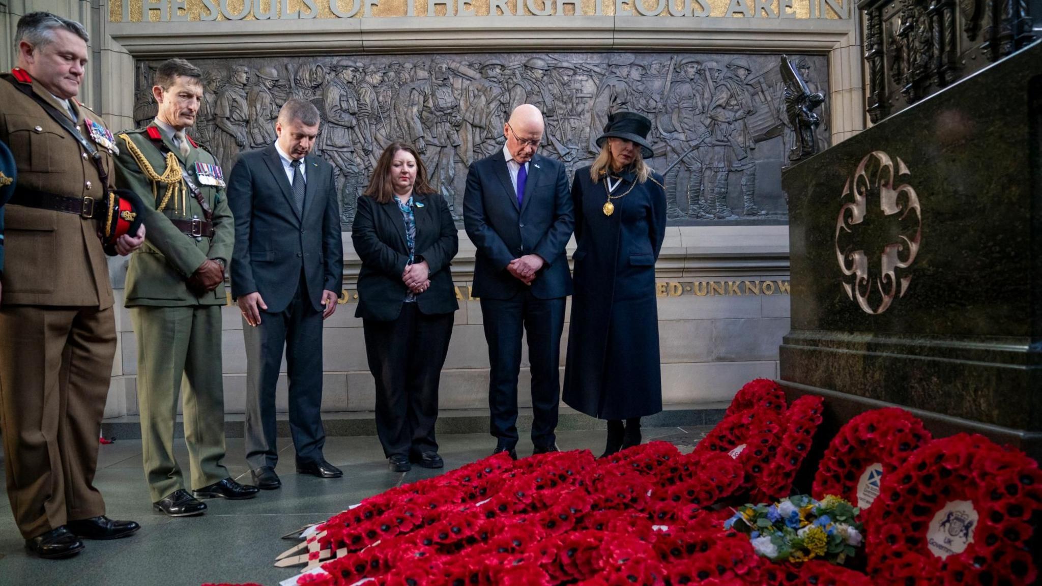 A number of dignitaries, some in uniform, look down at poppy wreaths at a war memorial