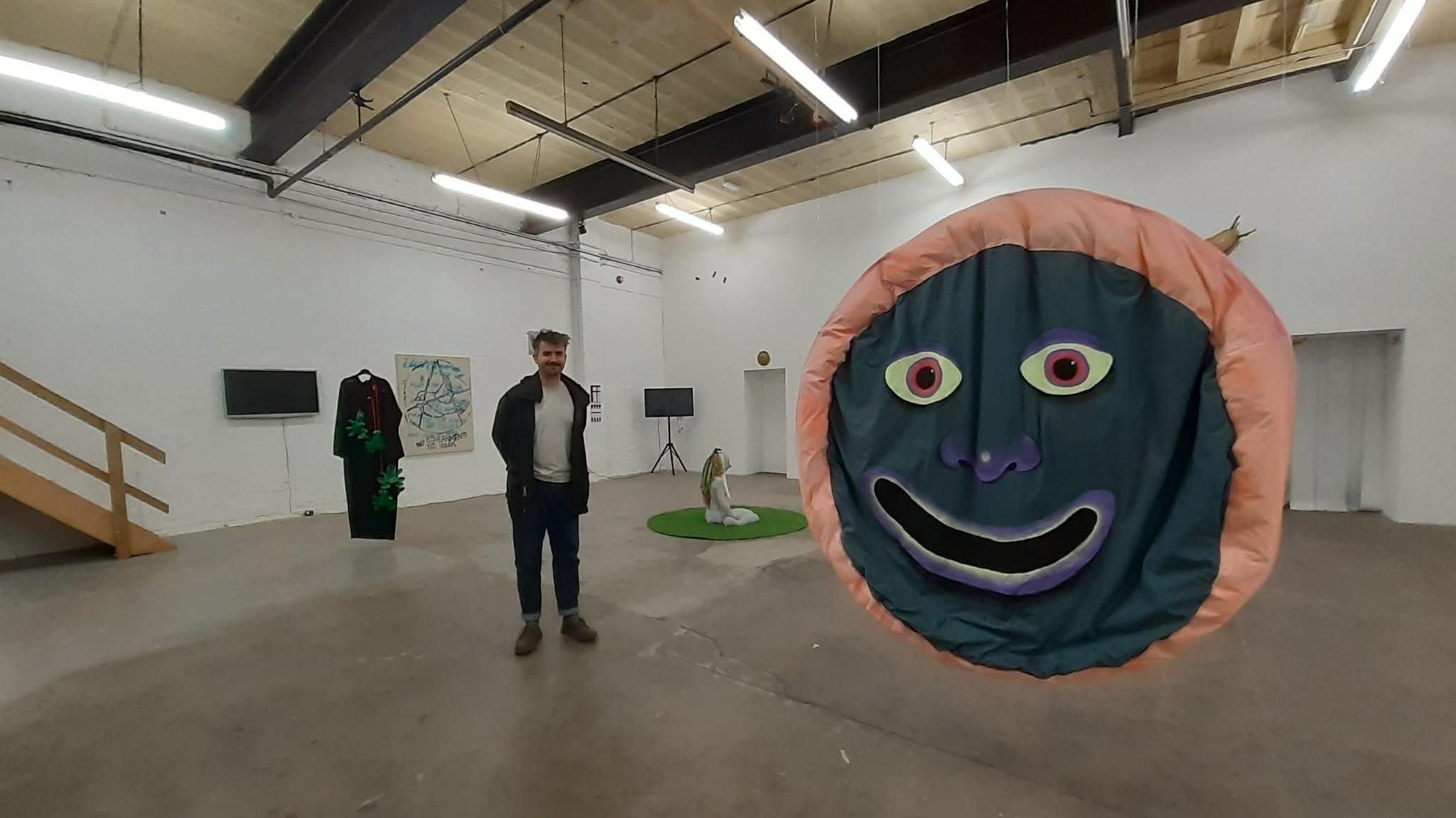 A man standing in a large white-washed room with paintings on the wall and a large smiling face art exhibit in the foreground