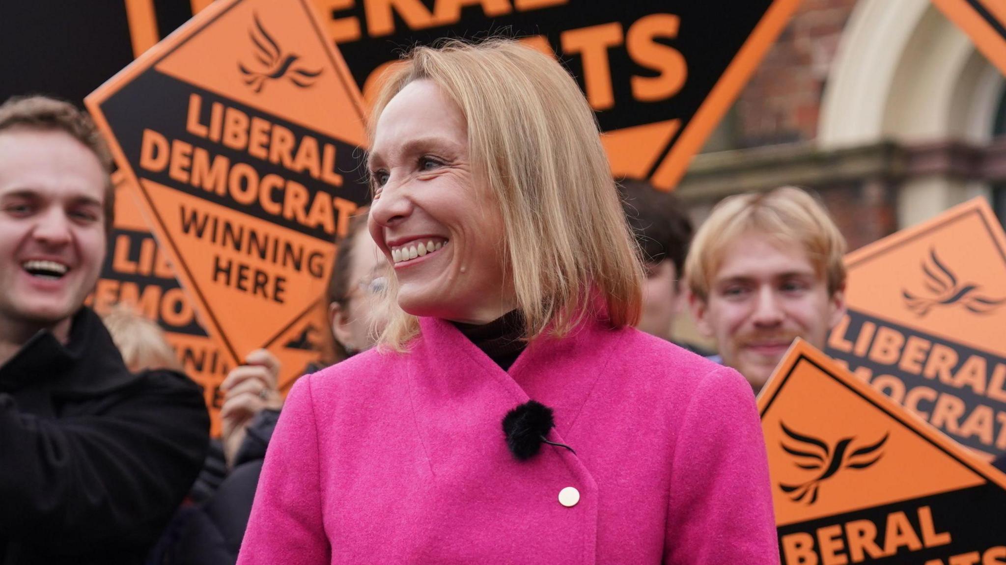 Helen Morgan, wearing a pink coat, smiling in front of Liberal Democrat campaigners holding orange placards 