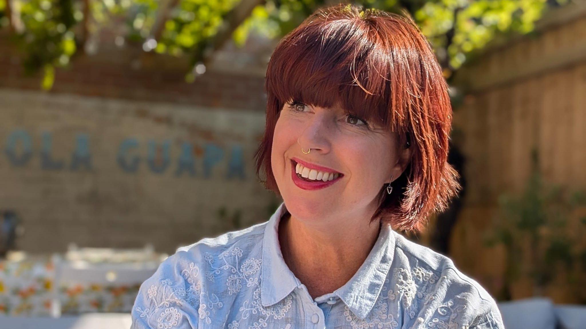 Catherine Warrilow smiles in a garden. She has red hair, nose jewellery and wears a blue top.