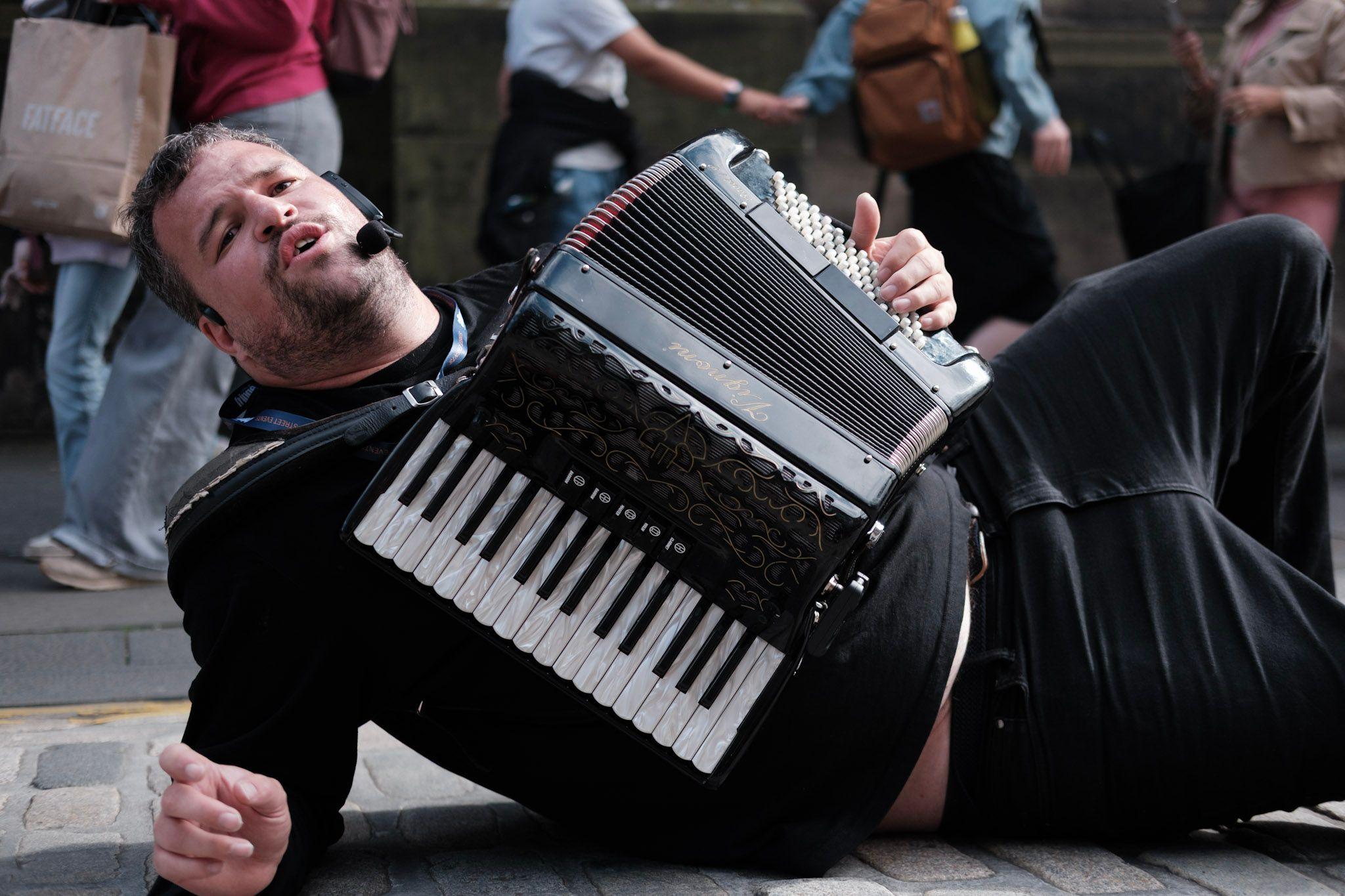 A bearded man in a black top and jeans plays the accordion as he lies on the cobbled street and sings. People are walking behind him.