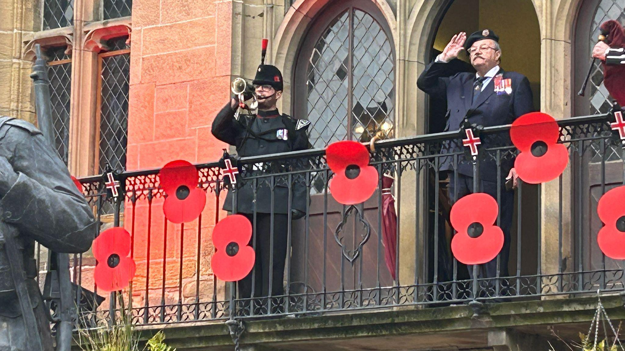 Arthur Lockyear, chairman of Durham City Remembrance Parade, is dressed in a military uniform and stands on a balcony decorated with large poppies at Durham Cathedral. A man also in uniform stands next to him playing the trumpet.