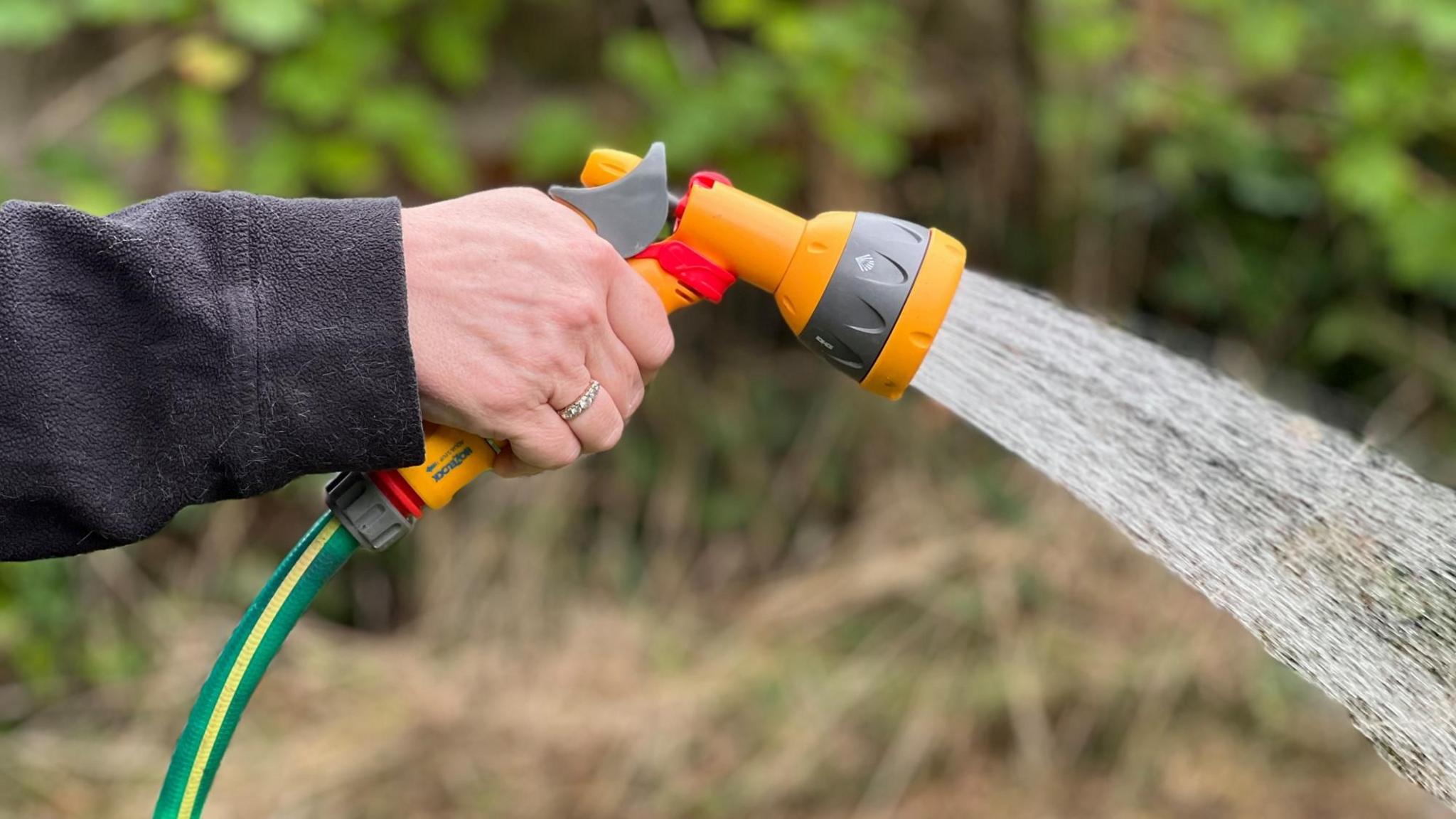 A hand holds an orange sprinkler with a green and yellow hose, which is spraying water out onto a garden.