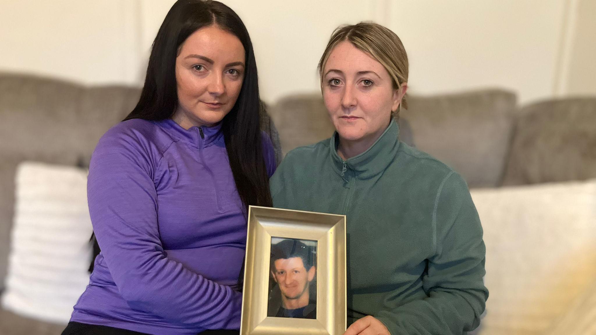 Danielle Bradley is in a purple zipped top and is sitting on a sofa.
Danielle has long straight, black hair. 
Ashling Graham is holding a picture of their father, Jim McFadden. 
She has short blonde hair.