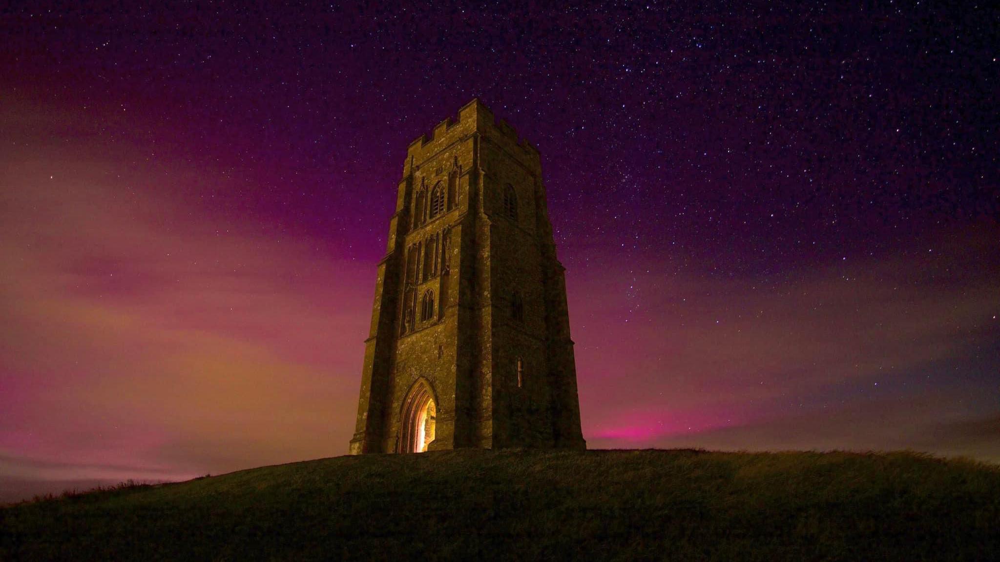 Northern Lights above Glastonbury Tor at night, causing purple streaks in the dark sky