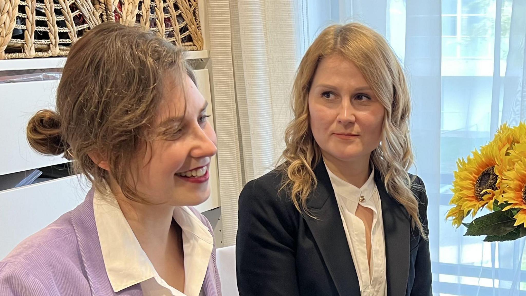 Two woman sit side by side next to a vase of sunflowers. They both wear white shirts with blazers - one blazer is purple the other black. 