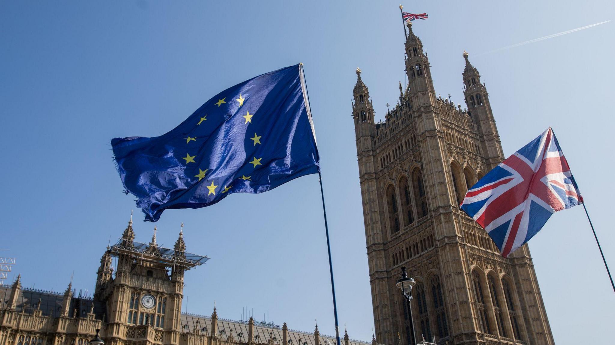 Houses of parliament in London with the EU flag and UK flag flying in front of it. It looks like a sunny day - the sky is bright blue.