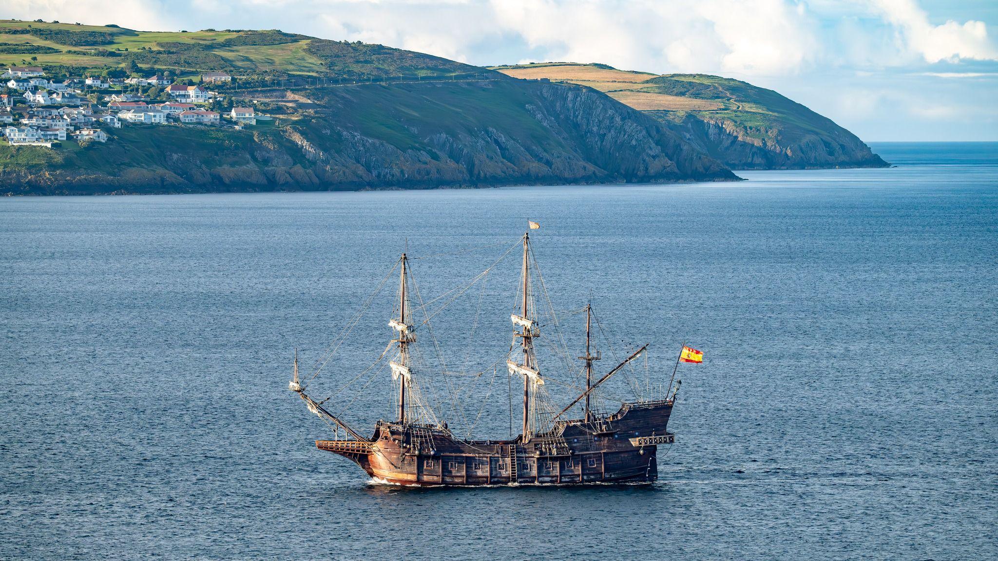 The large wooden vessel with three large masts, there is a Spanish flag on the bow. Onchan Head is in the background.