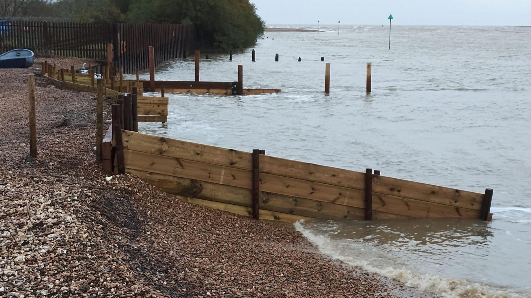 An image of the coastline, where the beach meets the sea. Wooden groynes and trees in the distance also feature in the image.