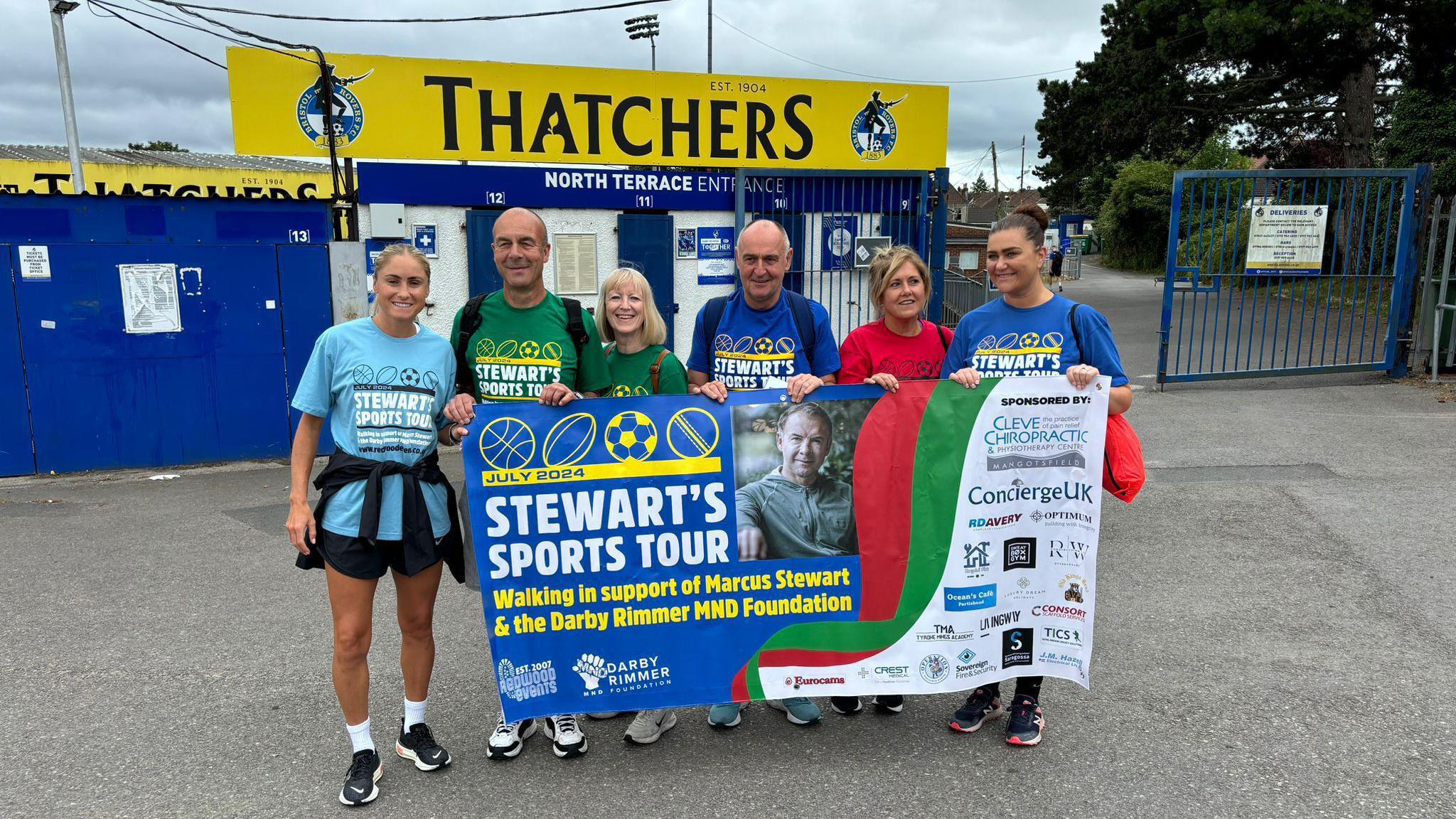 Six people holding a banner in front of a football stadium. The banner read 'Stewart's Sports Tour'