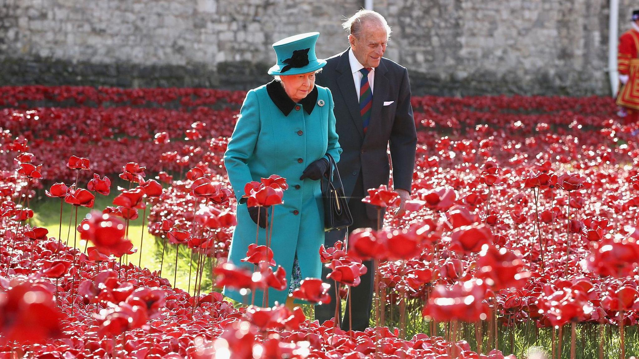 The Queen and Prince Philip walk among a sea of ceramic red poppies in the grounds of the Tower of London. The Queen is wearing a turquoise coat and matching hat. 