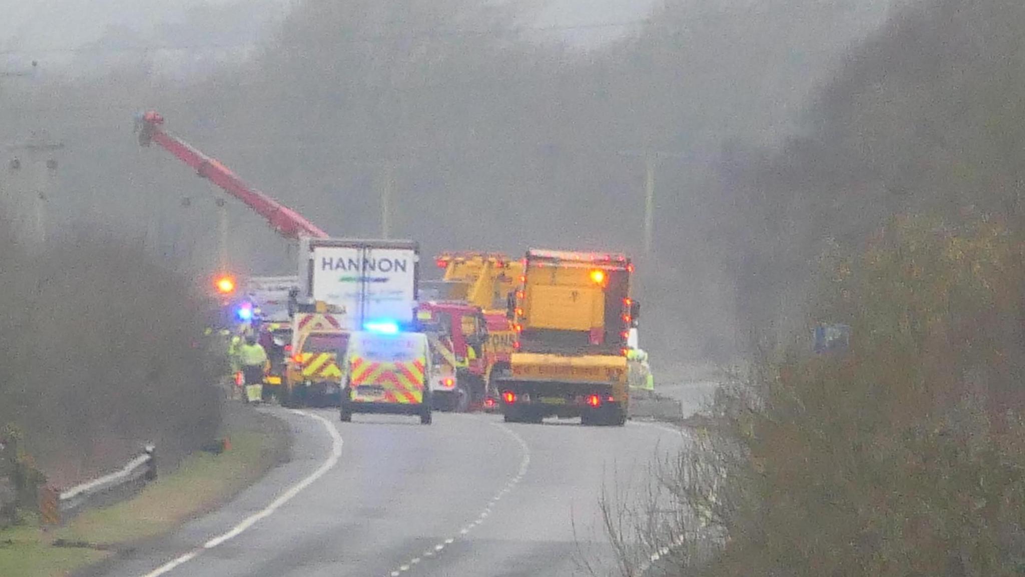 Emergency service vehicles and lorries block a road. It is a wet day. A man in hi-vis can also be seen