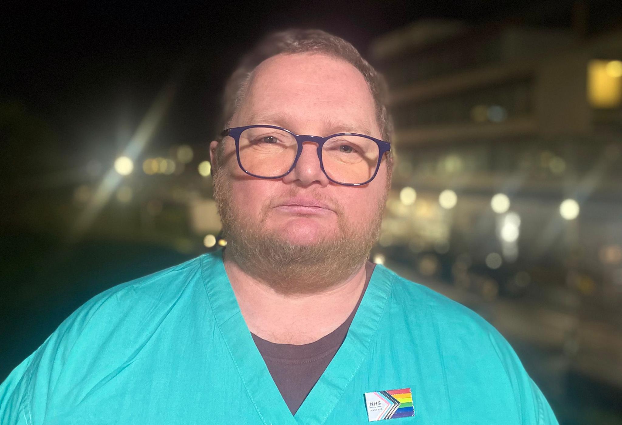 A bearded man with glasses and green medical scrubs stares directly at hte camera with the lights of a hospital building in the background