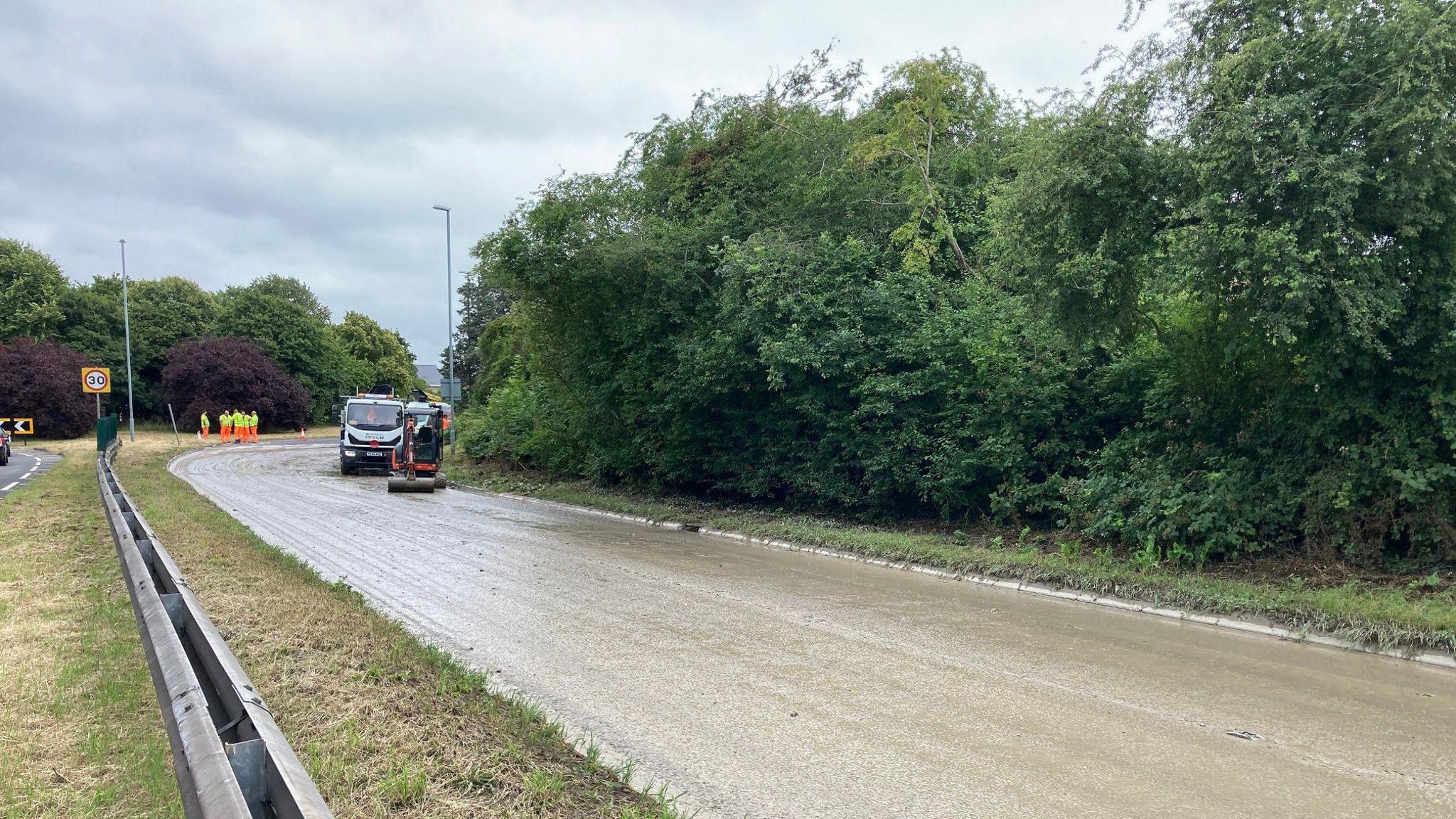 An alternative view of the road covered in mud, taken from the central reservation. Workmen can be seen standing in the distance, in front of a large roundabout.