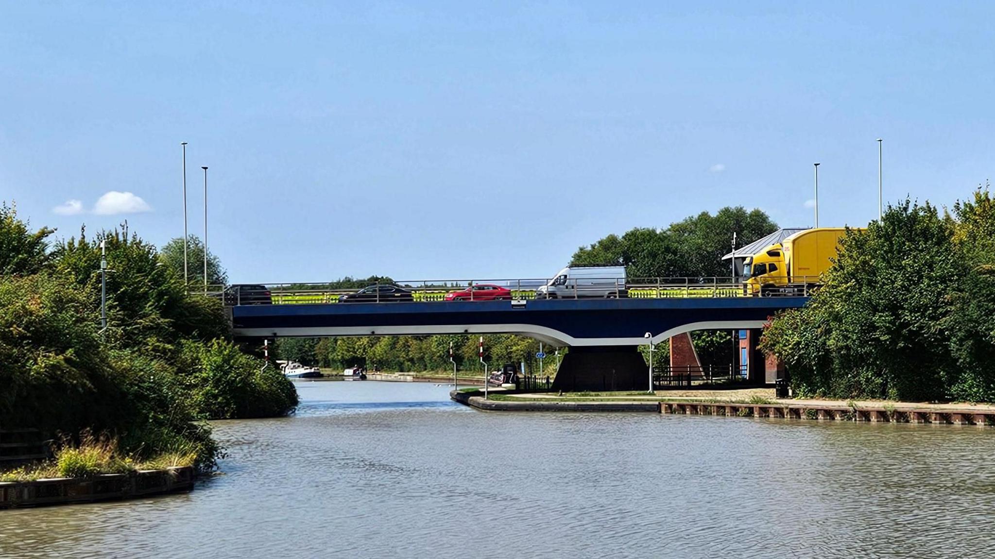 A bridge carries traffic, including a bright yellow lorry, a van, and three cars, over a canal on a sunny summer's day. There are trees on either side of the canal and canal boats can be seen in the distance on the other side of the bridge. There are hardly any clouds in the sky.