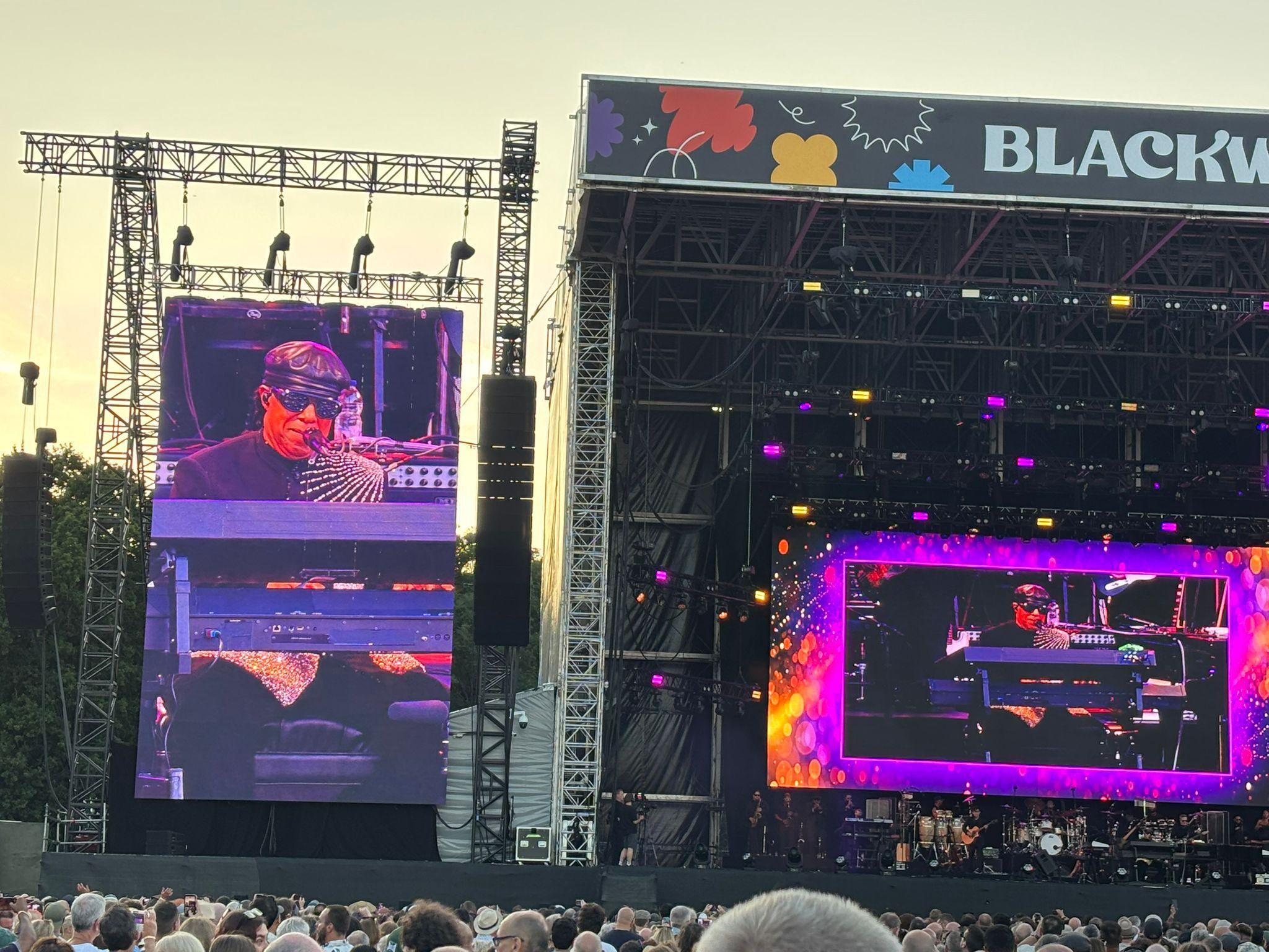 A shot from the crowd of the Stevie Wonder concert in Cardiff. His face, wearing a black hat and sunglasses are on a screen on the left of the shot and in the middle of the stage. The letters Blackw appear at the top of the stage 