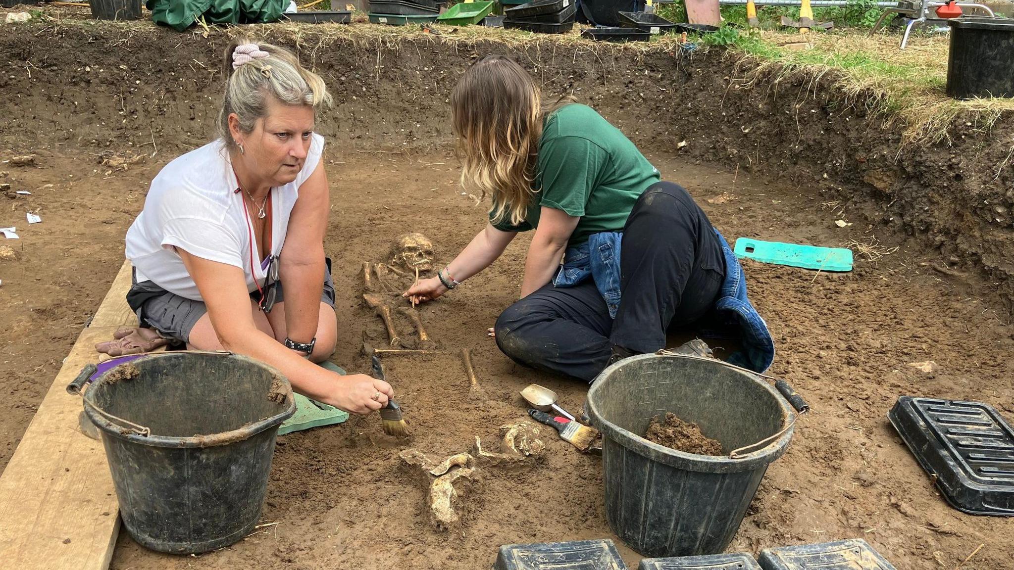 Two women, one with grey hair and wearing a white shirt and grey shorts, and the other with blonde hair and wearing a green top and black trousers, sitting on the floor of an archaeological pit. They're using brushes to brush soil away from a partially-excavated skeleton. They have buckets and tools on the ground around them.