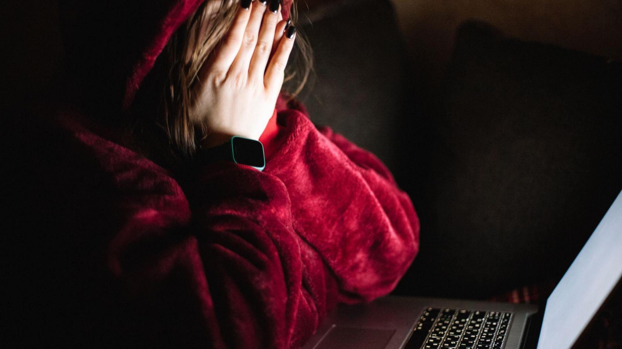 A person in a fluffy red jacket covering their face with their hands, which have black nail polish on them. They are sitting in front of a laptop which has a white screen.