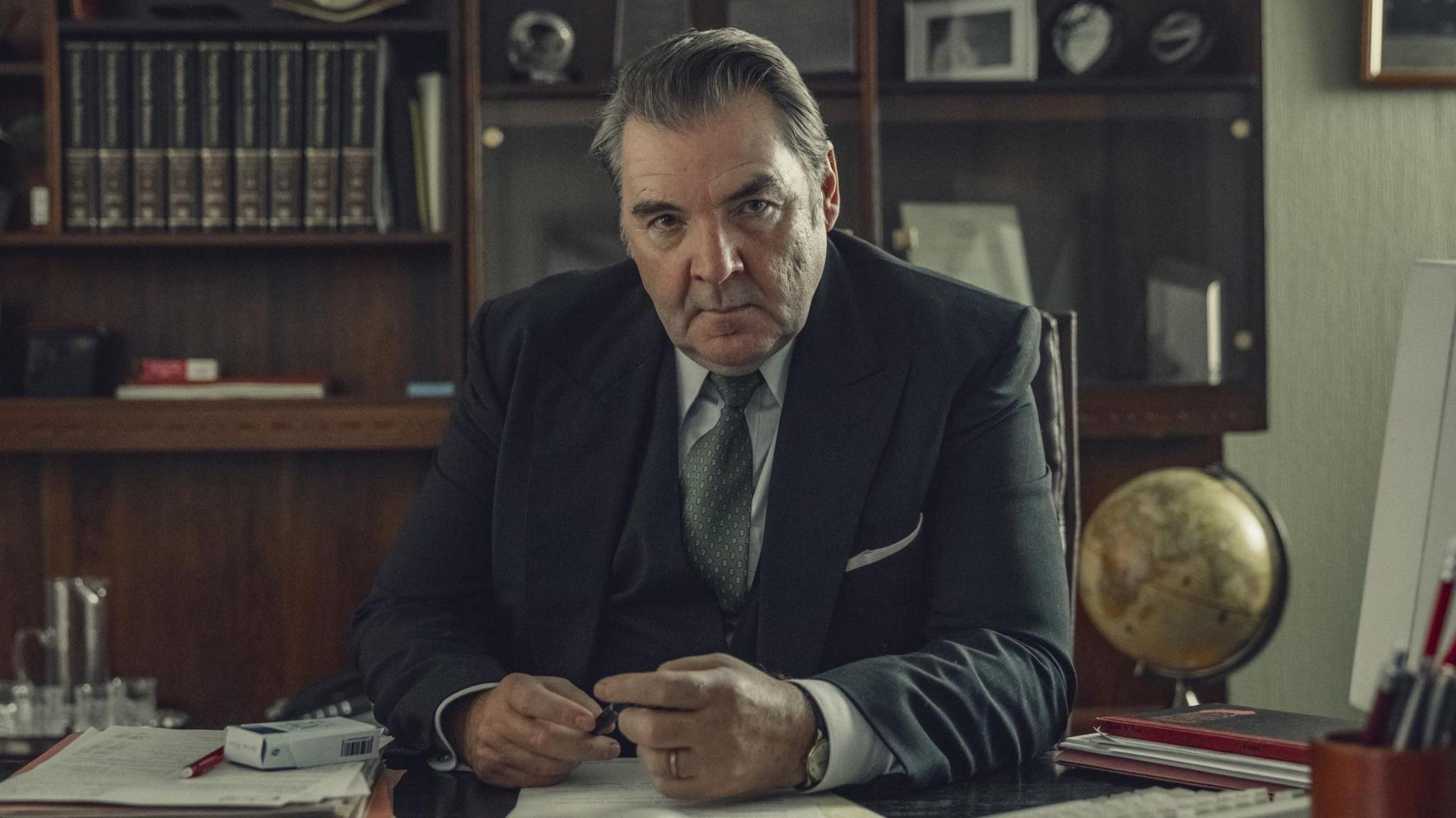 Brendan Coyle as Roy in Toxic Town. He is sitting at a desk with a bookcase behind him. He is wearing a black suit.