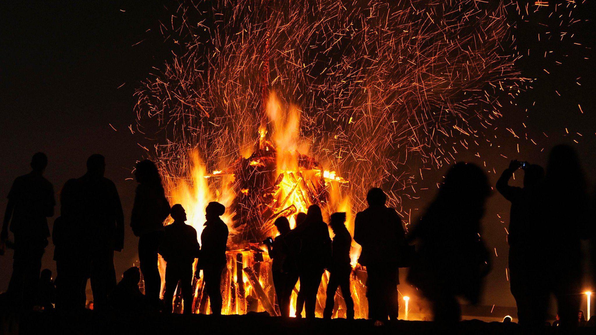 A group of people silhouetted at night against a large, blazing bonfire in front of them.