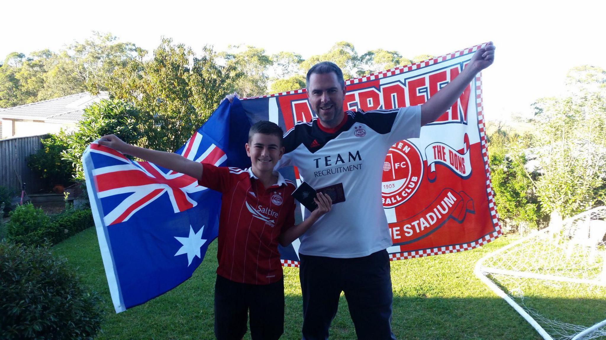 Jamie and Allan in 2017 before heading to Scotland for the cup final. Jamie is holding an Australia flag, which is a blue background with white stars, and a red, white and blue Union Jack style flag in the corner. He is wearing a red Aberdeen FC jersey. Allan is holding a red flag which says "Aberdeen" and has a blue and white saltire cross on one side. He is wearing a white and black Aberdeen jersey. They are standing in a garden.