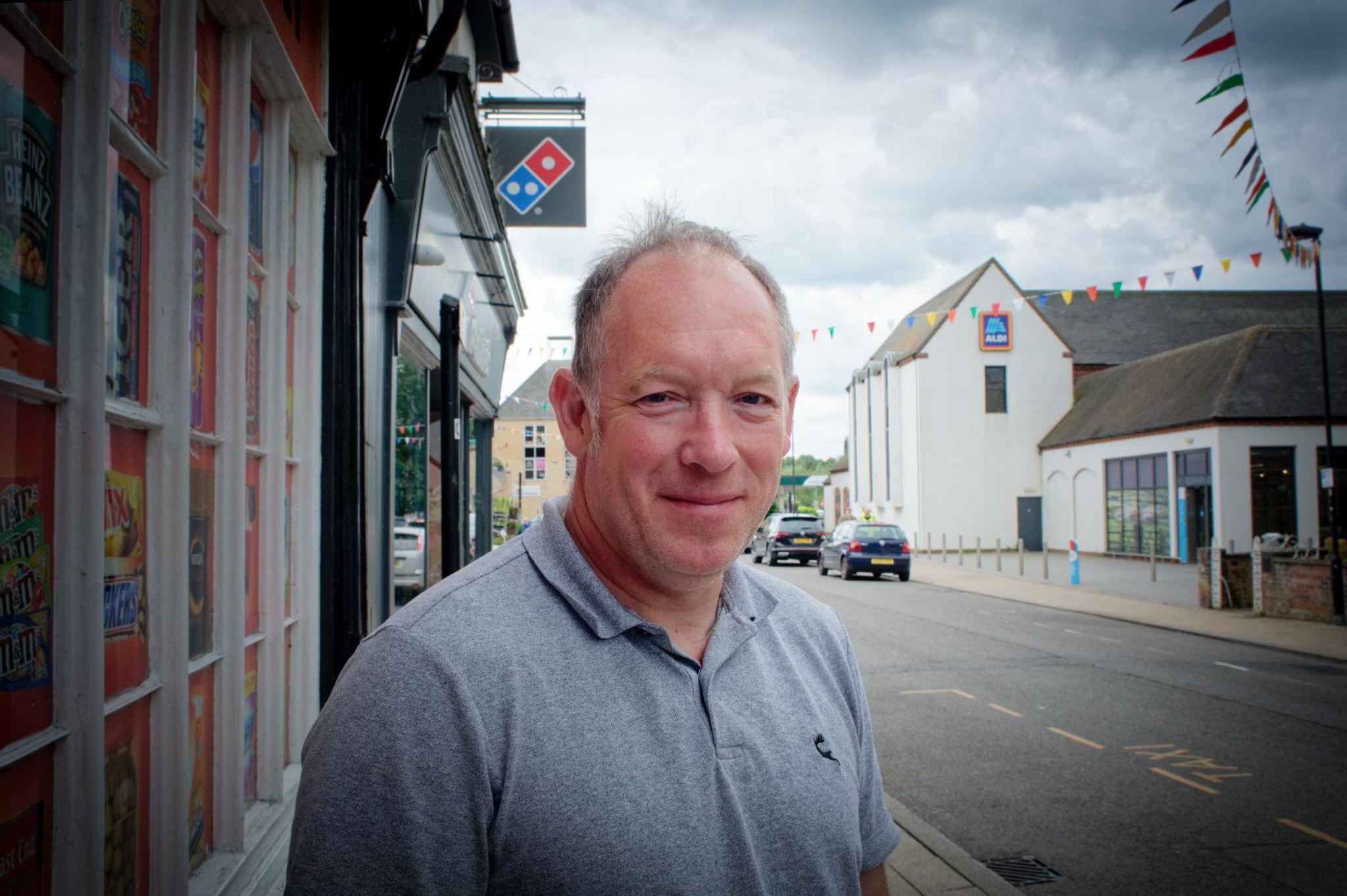 Peter Banham-Gibbs is smiling in the street. There is bunting behind him