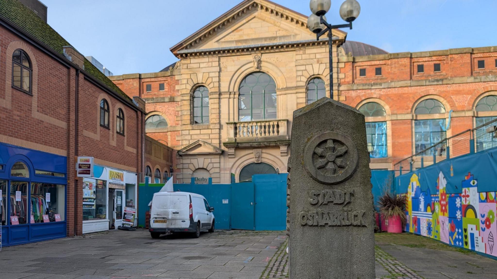 A stone monument in front of Derby's market hall. Behind the stone and in front of the market hall is blue temporary fencing.  