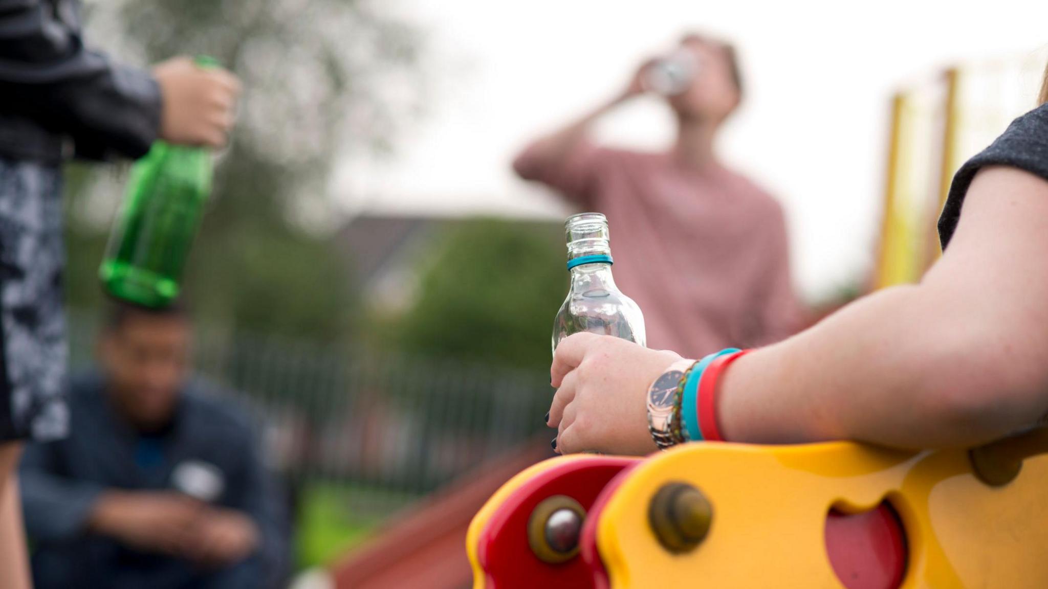 Teens drinking in a park in daylight, images blurred. Four figures can be seen, holding and drinking from bottles which appear to hold alcohol. Yellow and red play equipment can be seen and green railings around a children's play area.