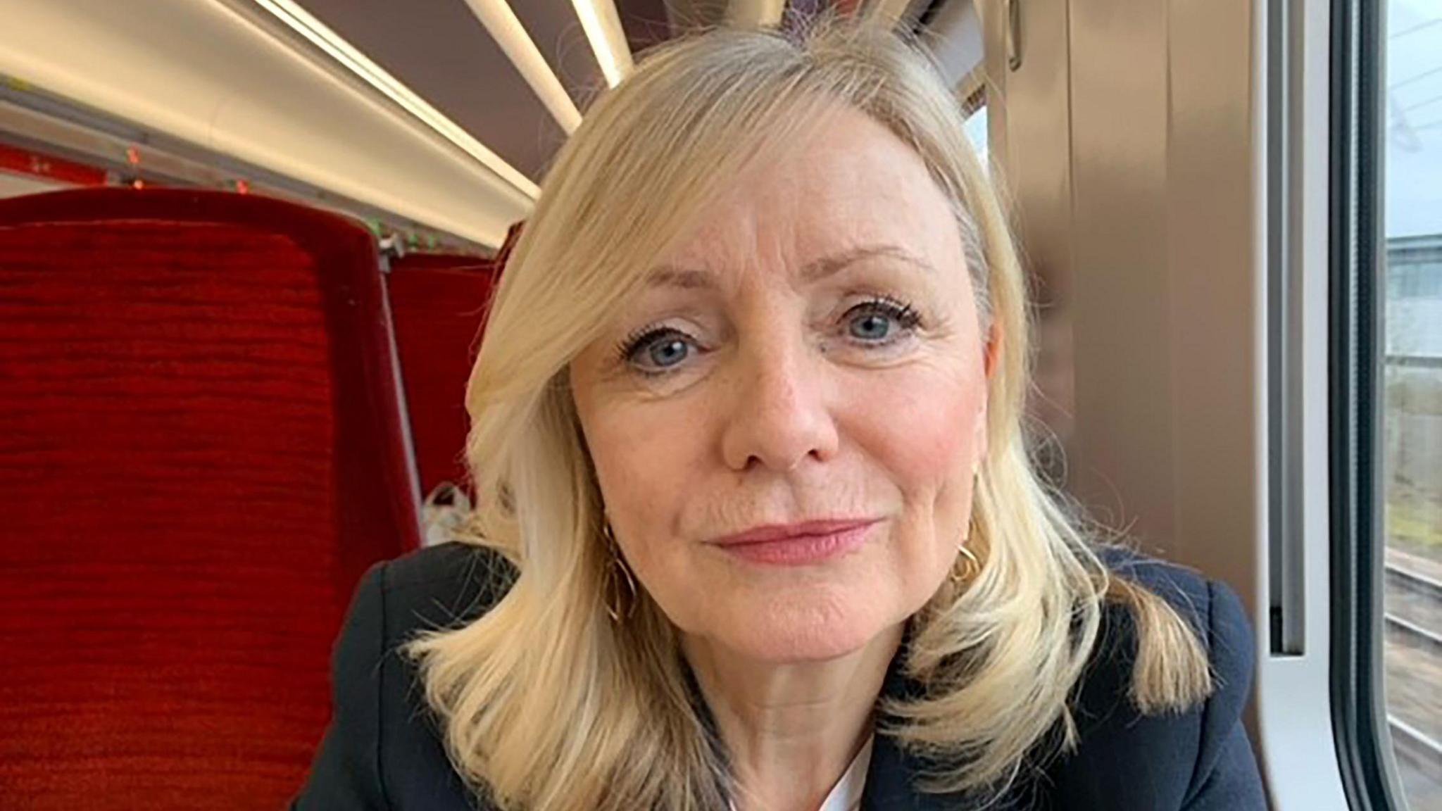 Tracy Brabin looks into the camera. She has long blonde hair and is wearing a navy jacket. She is sitting on a train seat.