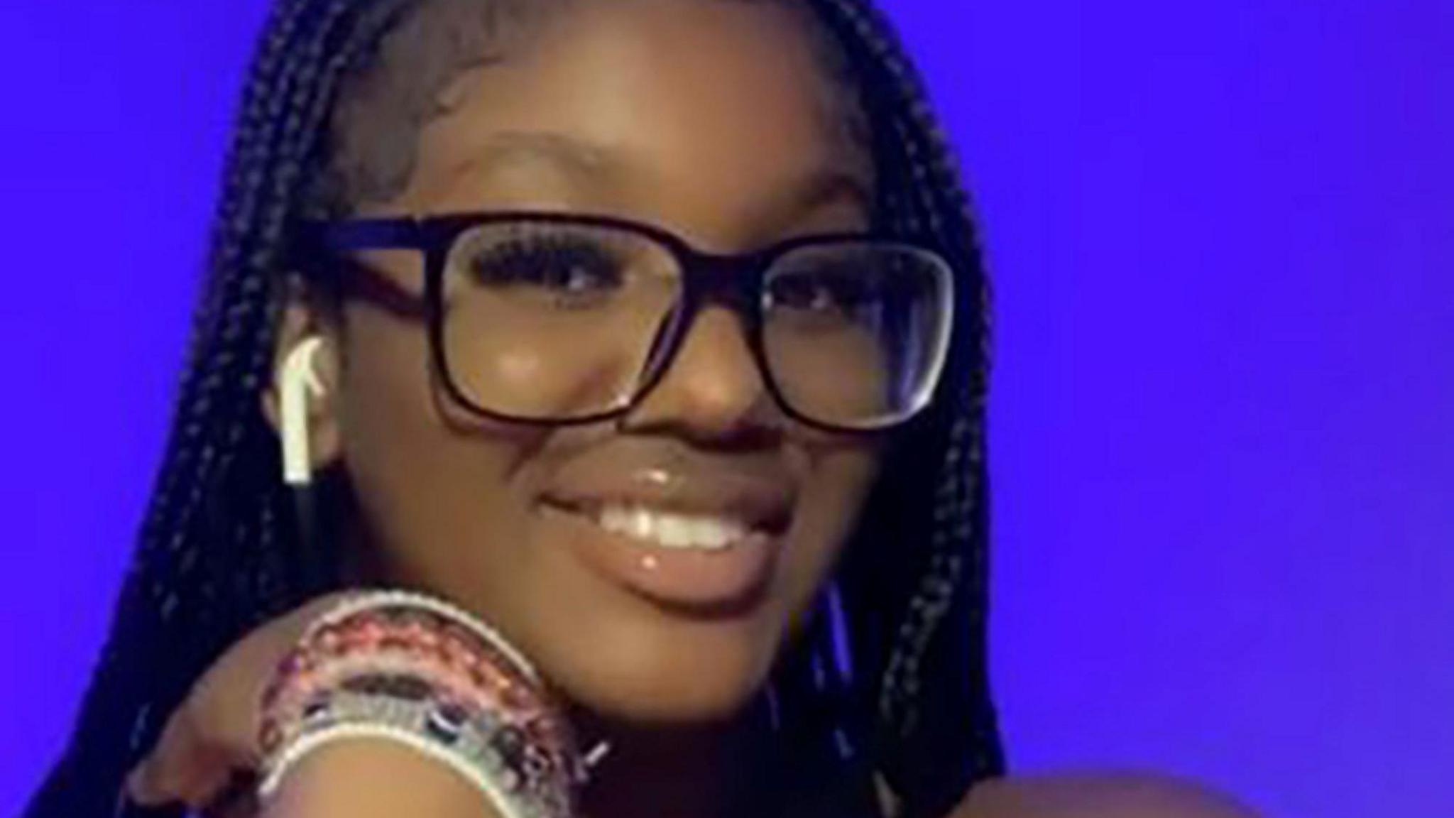 Elianne Andam headshot smiling at the camera with a bright purple background. She wears a number of bracelets on one wrist, black-framed glasses and one earphone.