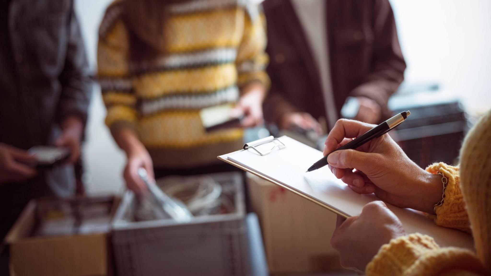 A person is writing on a clipboard in the foreground. in the background, three unidentifiable people sort through boxes