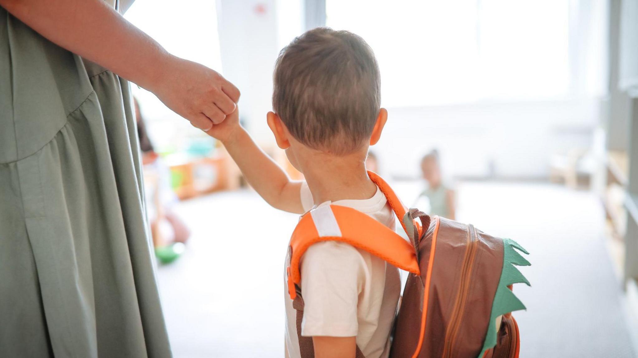 A young boy with an orange backpack on holding his mother's hand and looking into the distance.