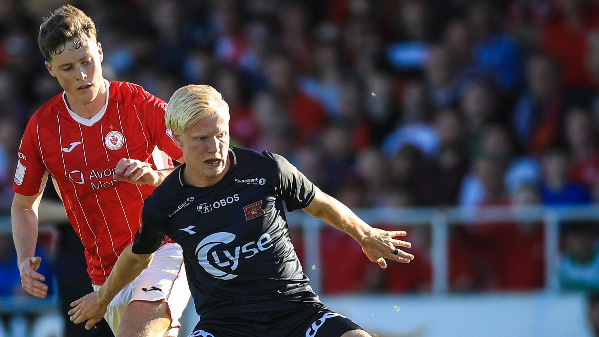 Harald Nilsen Tangen is pictured in a black playing shirt, holding off an opposition player who wears red. The background is blurred, but it is full of fans. 