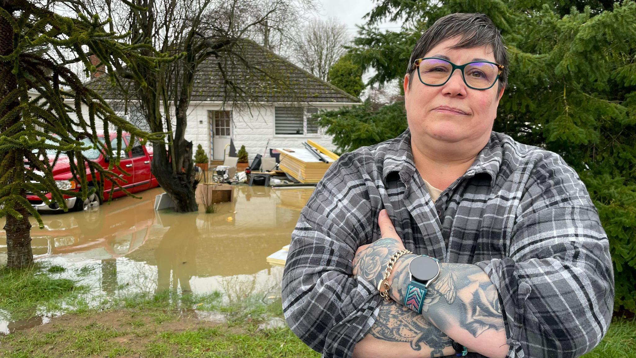 A woman with short black hair and glasses in a chequered shirt who has tattoos on her arms crossing them and looking unapprovingly into the camera. In the background you can see the front garden of a bungalow flooded with a red car submerged in it.