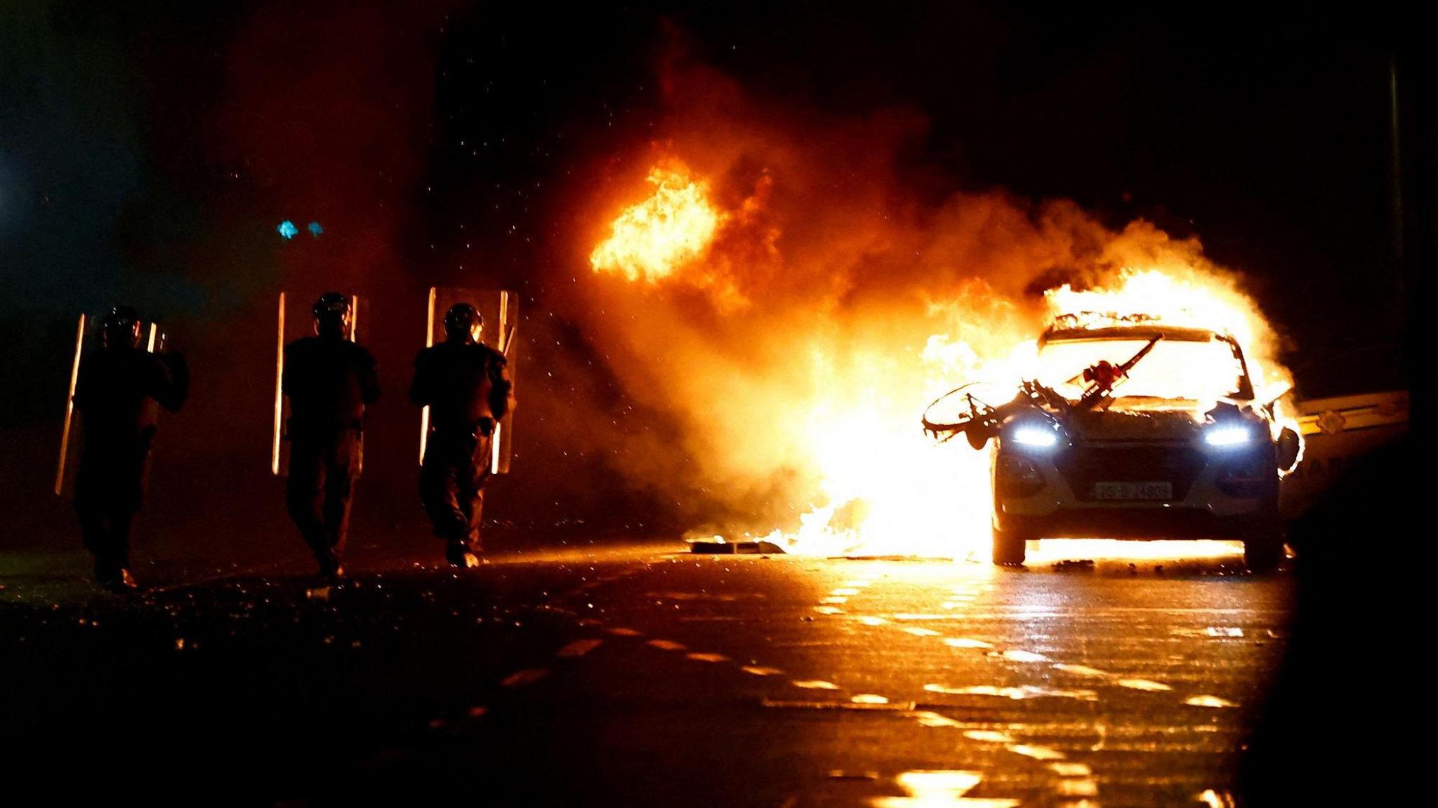 Riot police officers face down demonstrators next to a burning police car 