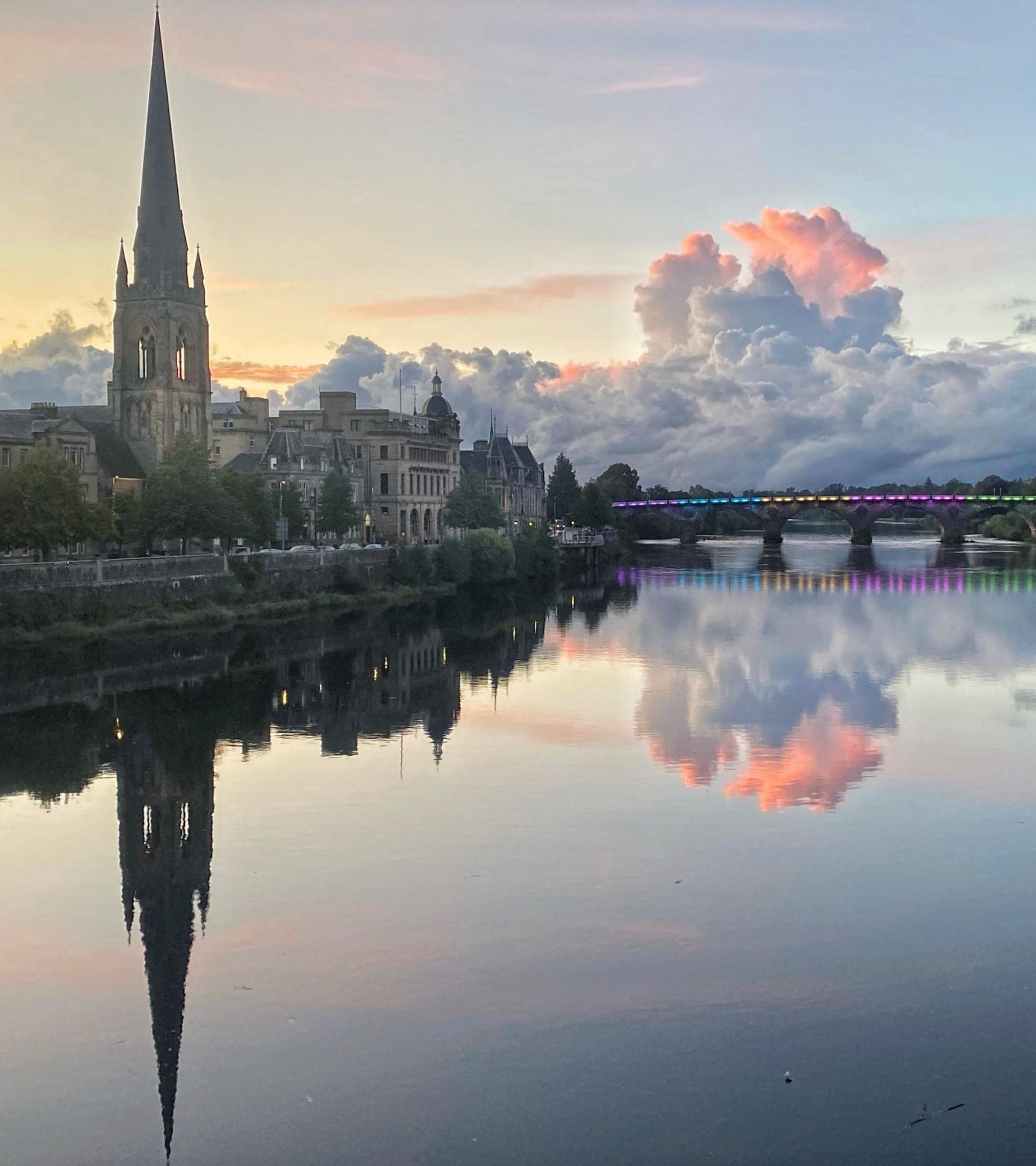 A view of Perth from across the Tay in the evening - the sky is a light blue with clouds tinged in a pink light. The bridge in the distance is lit in pastel colours adding to the magical feel