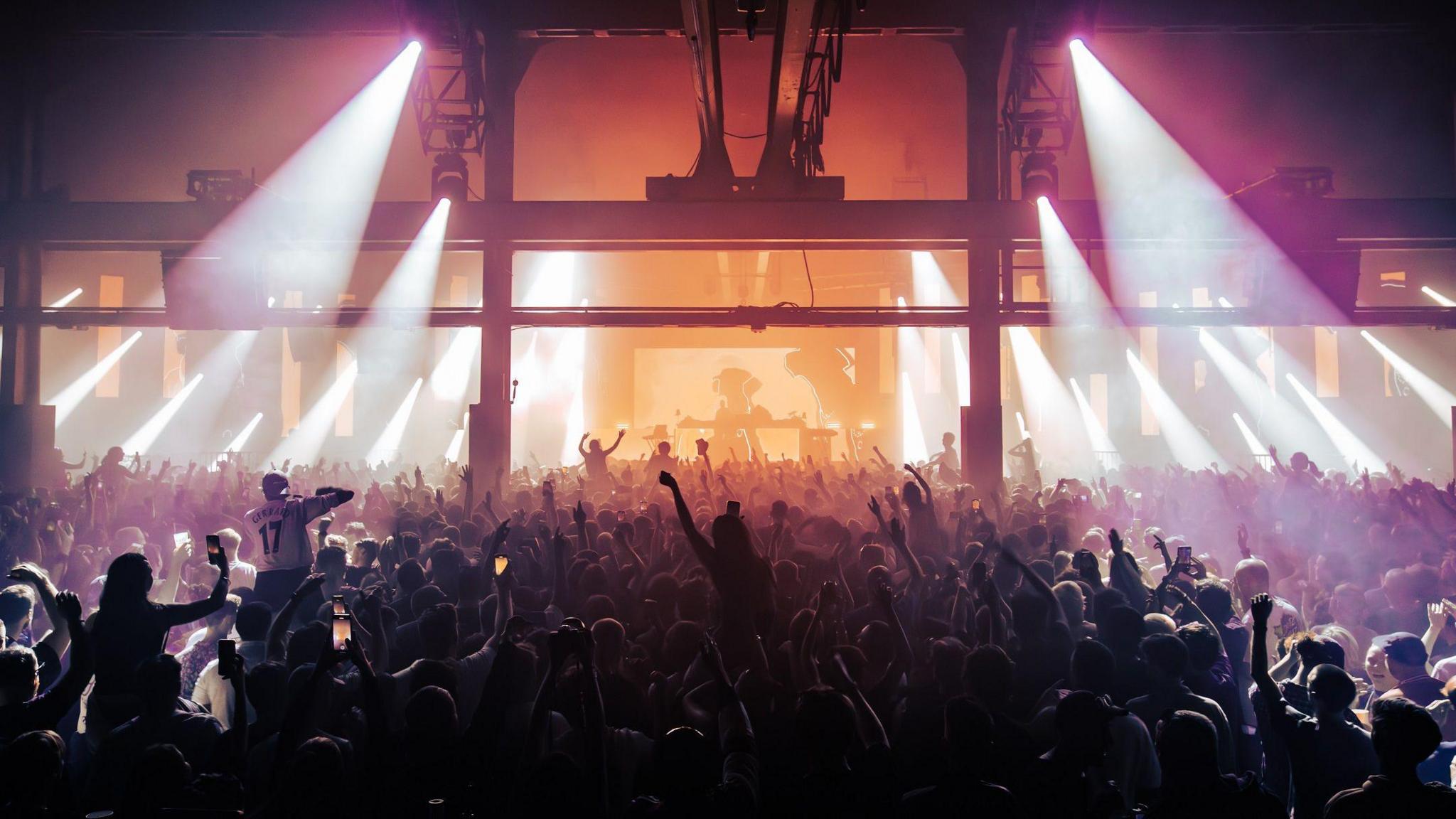 A large warehouse venue called Prospect in Bristol is packed as people dance during a DJ set. Some people in the crowd are on the shoulders of others and above them is industrial machinery including a huge internal crane.