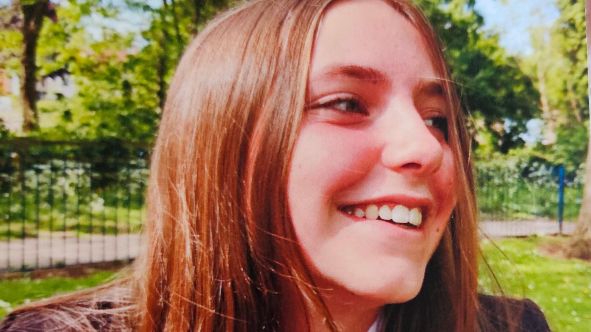 Chloe Longster wearing her high school uniform in a park. She is smiling and her head is turned to the side, she is looking to someone else out of frame to the side. She has long, straight brown hair and it is a sunny day