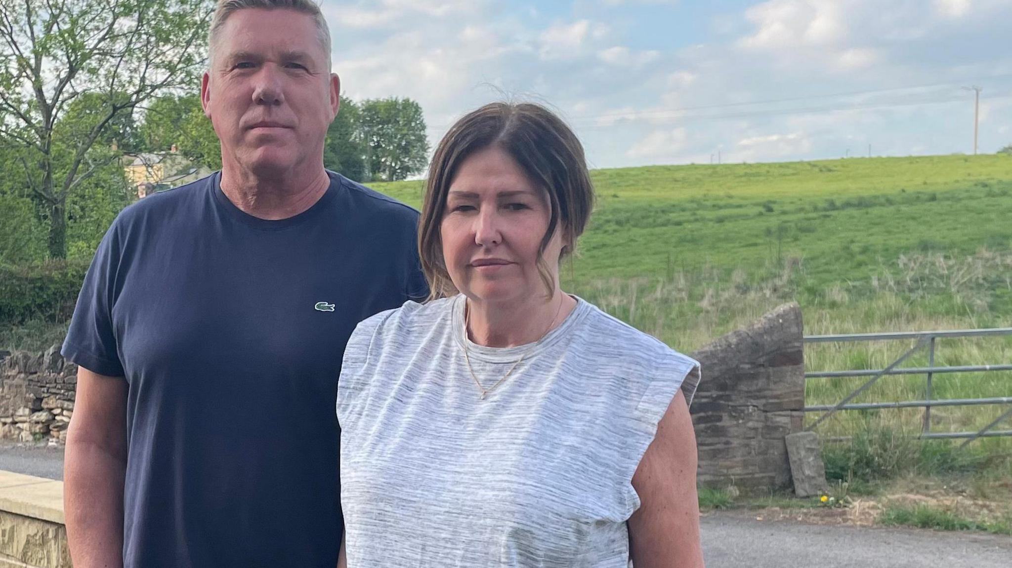 A man in a navy blue t-shirt stands next to a woman with dark hair wearing a grey t-shirt. Behind them is a wall and a gate, with an open field beyond.