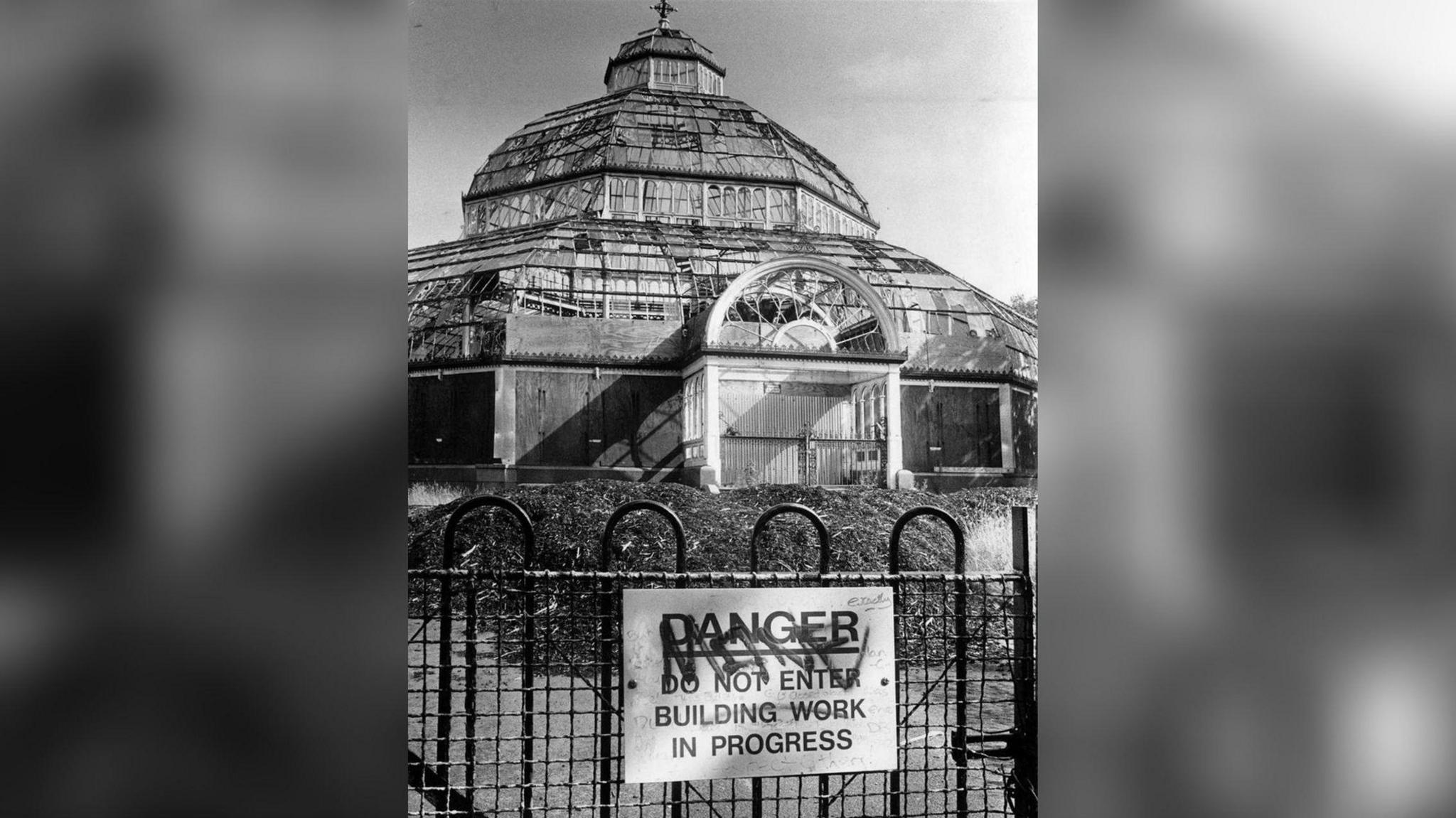A black-and-white photo shows a sign saying 'do not enter - building work in progress' on a barrier in front of the Palm House. It has much broken glass and missing glass in its frame.