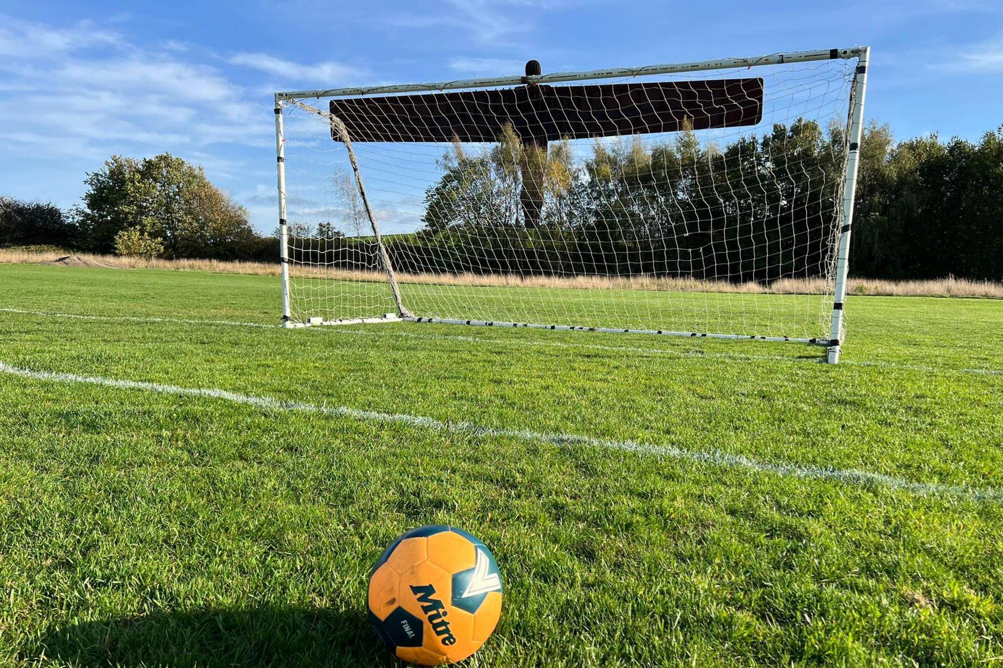 A goal on a grassy football pitch with an orange football placed in front. The Angel of the North sculpture looms overhead in the distance.