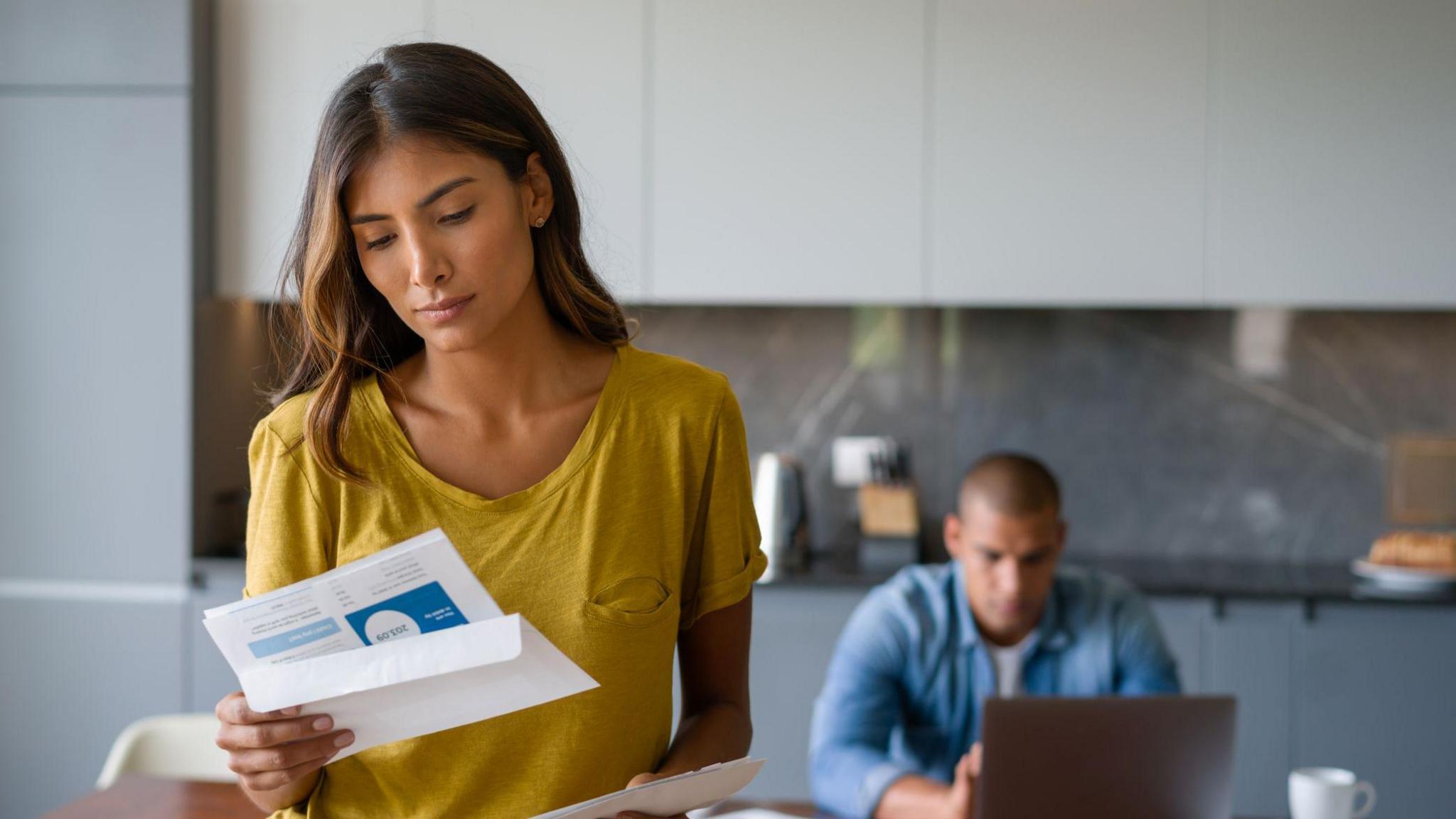 Woman looking at a bill in the kitchen of a home with a man sitting in front of a laptop in the background.