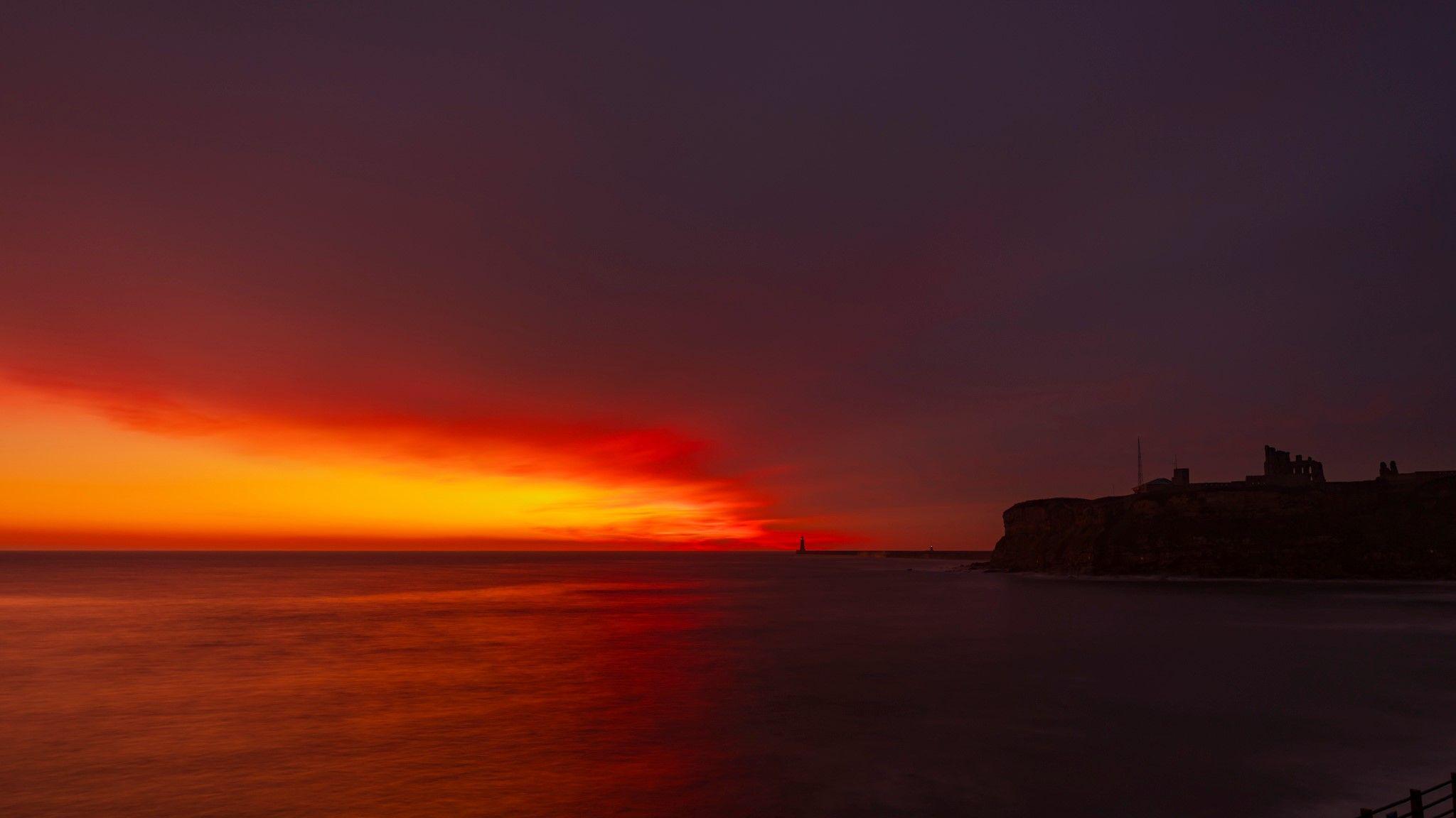 Looking out to sea at Tynemouth. The sky is largely dark but to the left there is a bright orange and red patch which looks to be spreading further right. The priory is to the right of the photo and is completely in shadow. In the distance the pier and lighthouse can be seen.