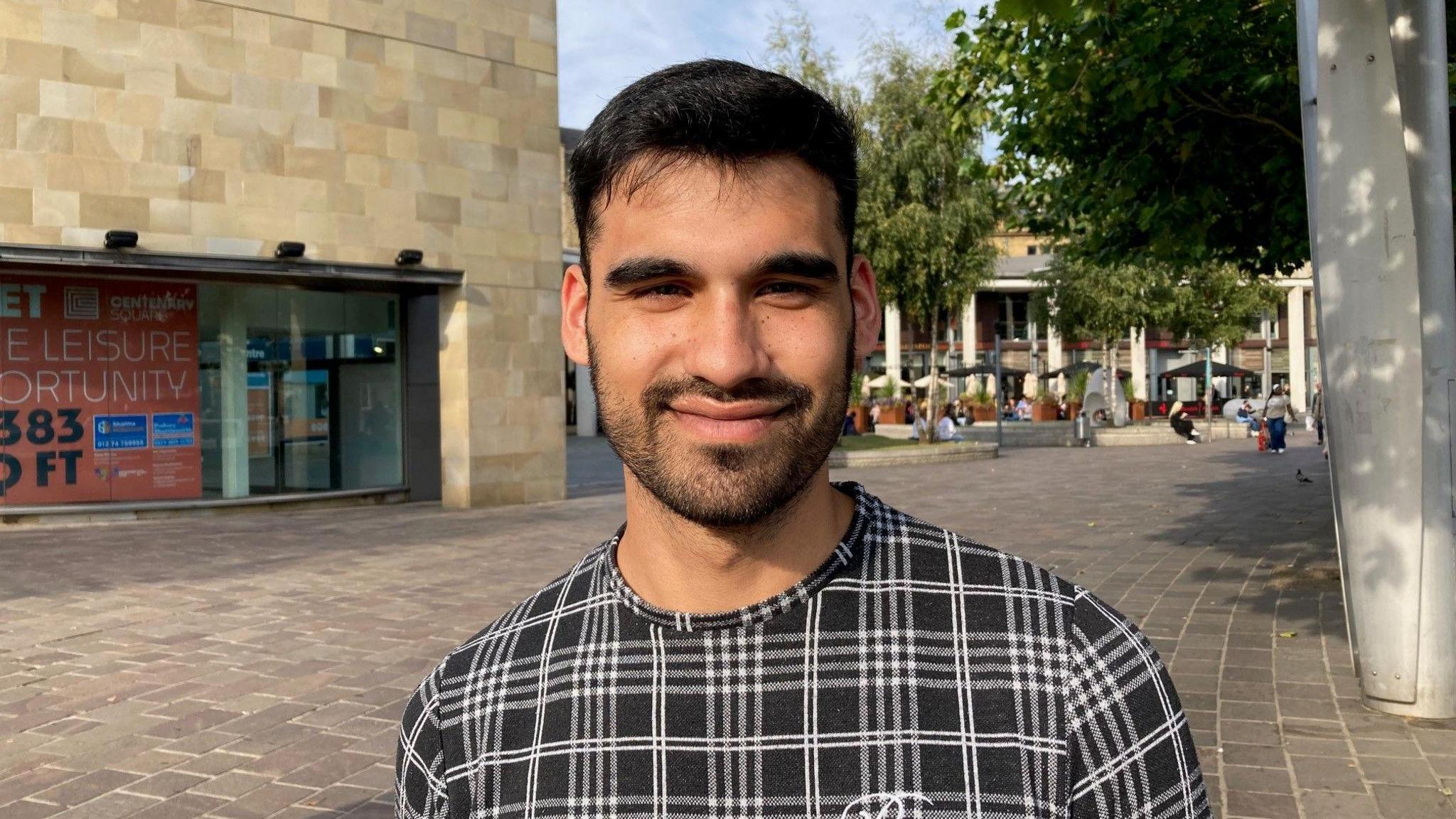 A brown-skinned male with short, dark hair and dark facial hair wearing a black and white patterned jumper looks at the camera