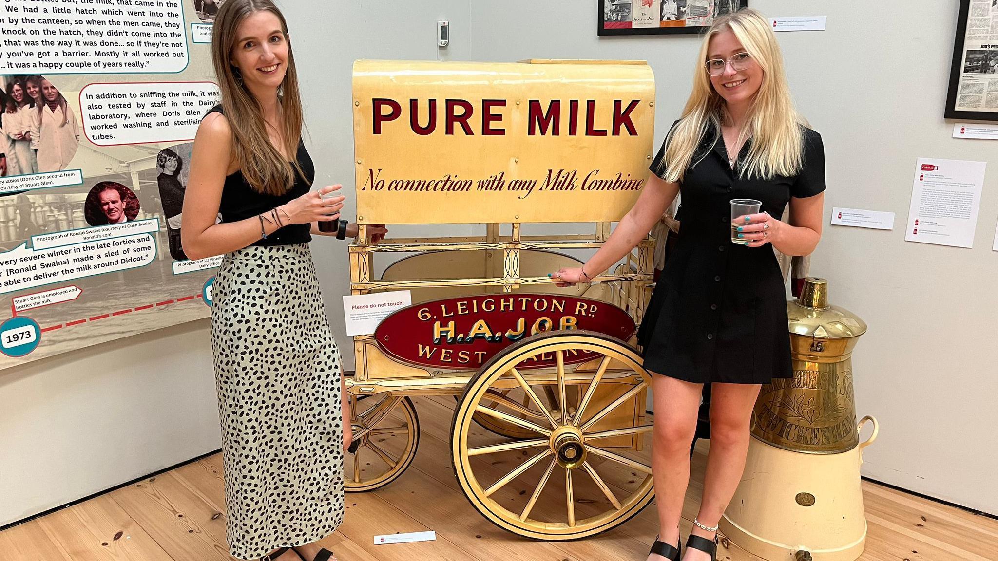 Ruby Livesey and Dr Leah Hewerdine are smiling at the camera, in front of a yellow milk cart which is part of the dairy exhibition. More memorabilia could be seen on the wall behind them. They are holding cups of drinks that were served at the event's launch.