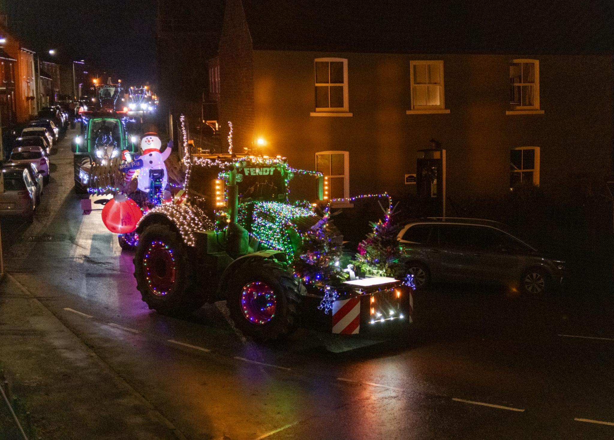 A tractor covered in bright Christmas lights travels through a village - there are more tractors following with an inflatable snowman and more lights on.