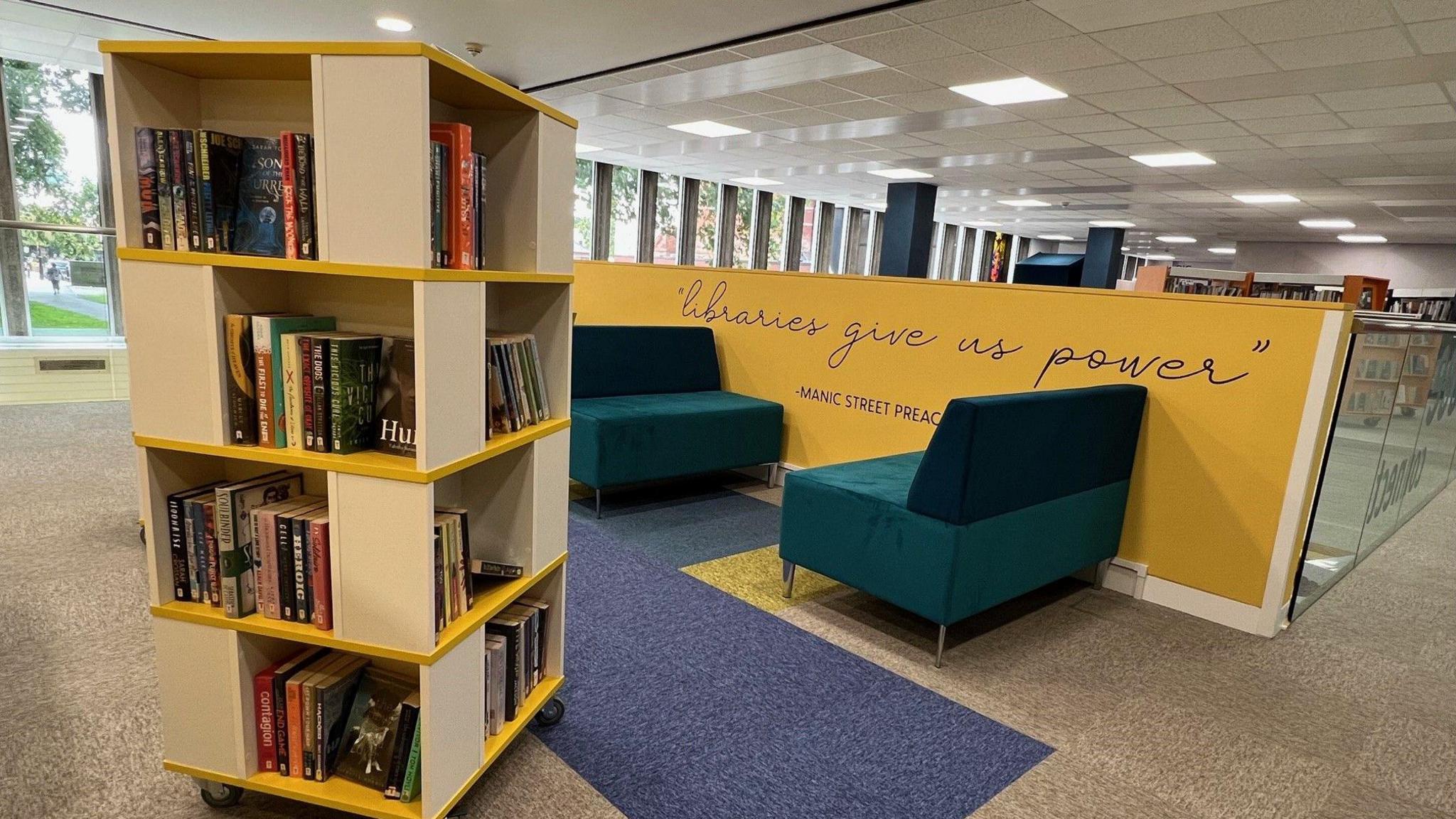 A white and yellow book shelf in a library, behind which is green cushioned seating positioned against a yellow balustrade. 