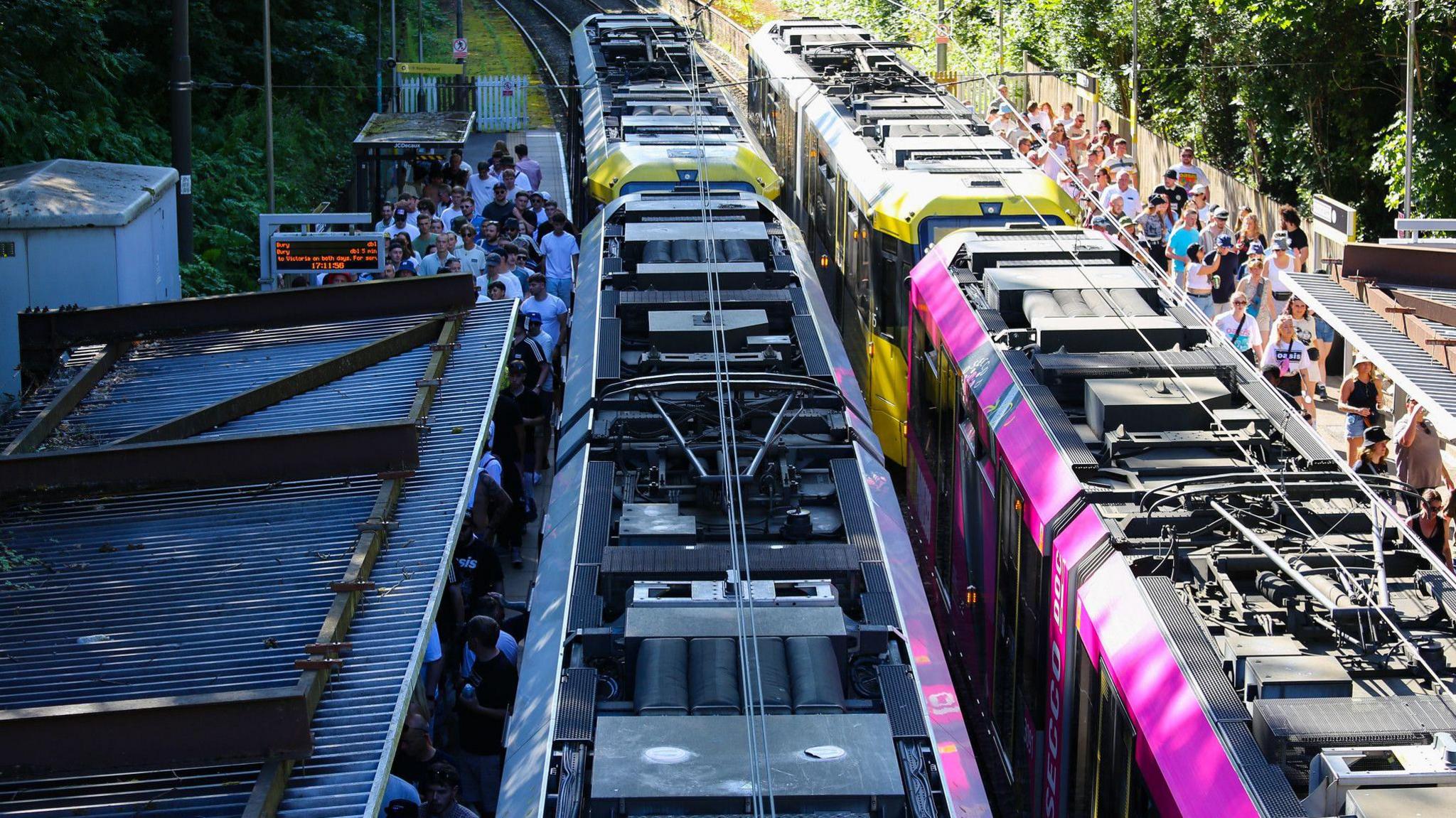 A long queue of Oasis fans stand under the corrugated iron roof of a Metrolink platform as a yellow and black liveried tram and a pink liveried tram stand at the stop